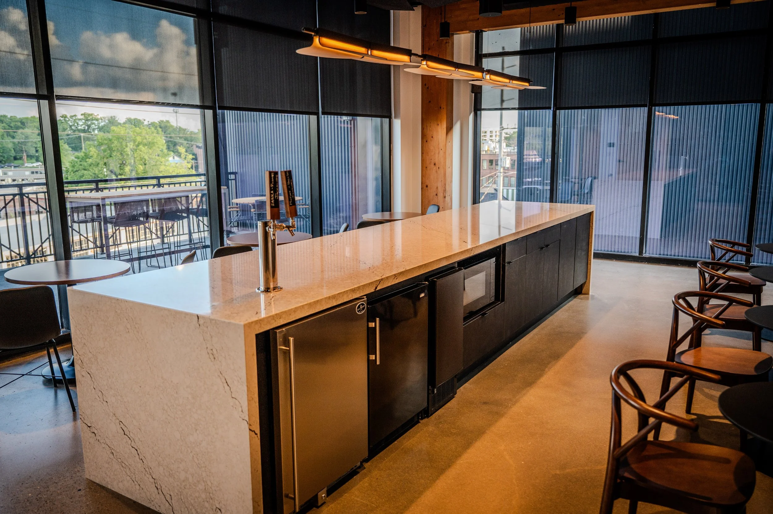 A modern bar/restaurant interior with a large kitchen island, black cabinets, wooden chairs, large floor-to-ceiling windows showing outdoor trees and sky, and a wooden ceiling with pendant lighting.