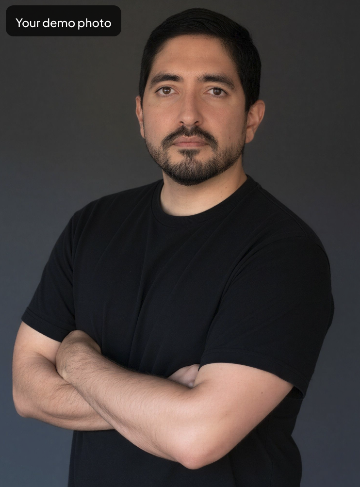 Portrait of a man with dark hair and a beard, wearing a black T-shirt, standing with arms crossed against a dark gray background.