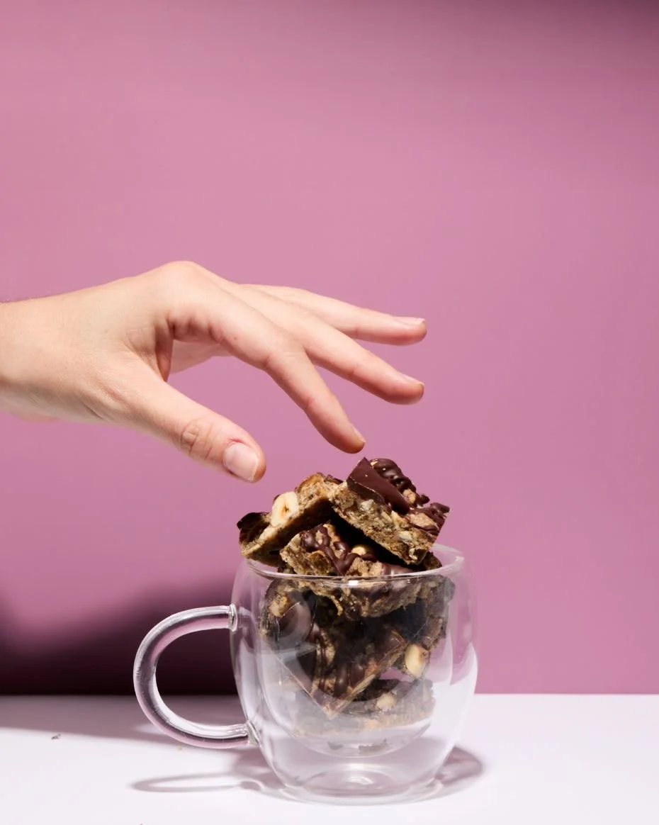 Hand reaching into a glass cup filled with chocolate chip cookie bars against a pink background.