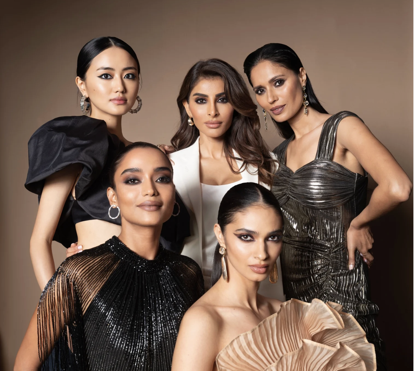 Group of six women wearing glamorous dresses, posing against a beige background.