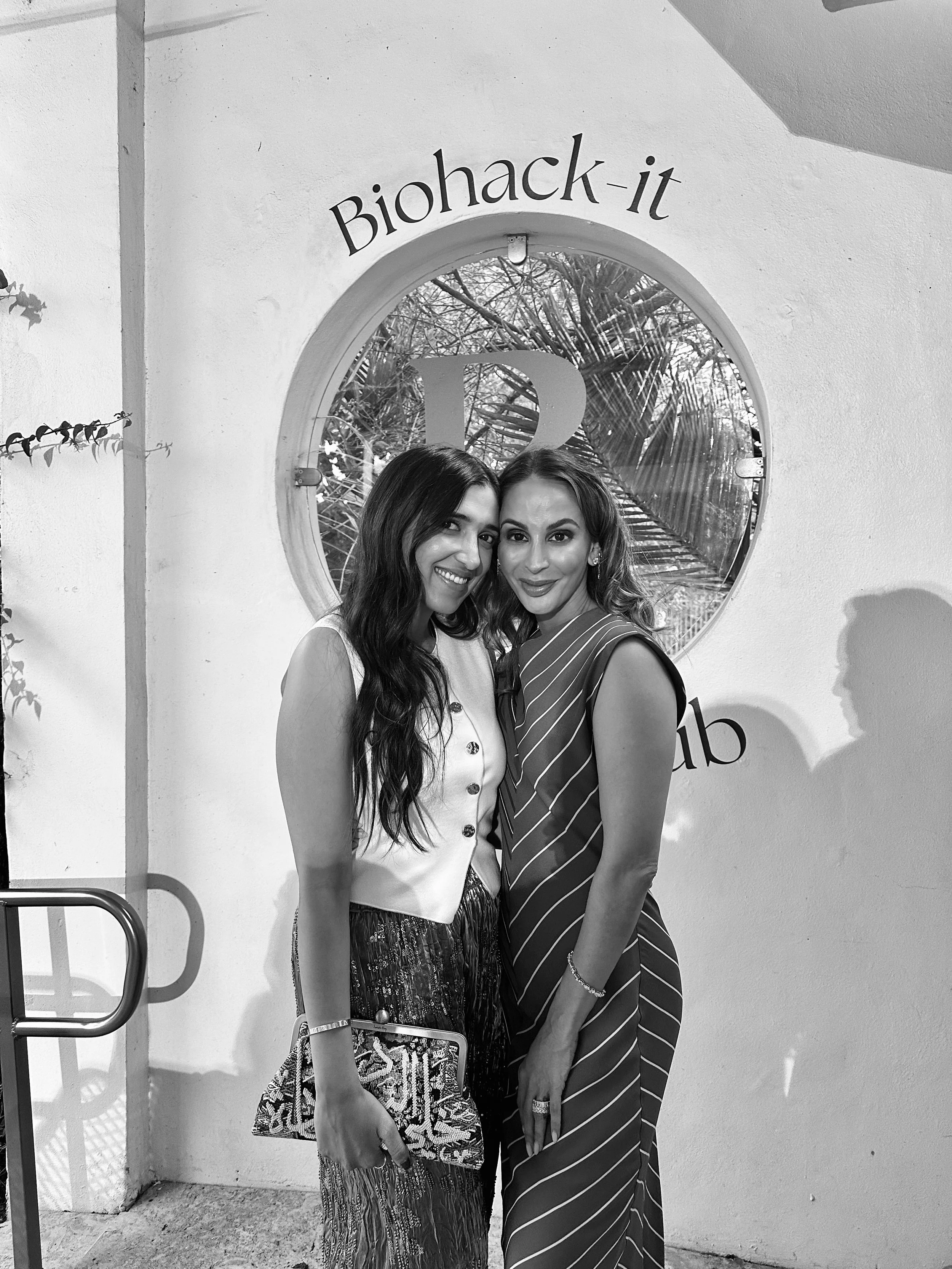 Two women standing together, smiling, at a location with a sign that reads 'Biohack-it' and a round window behind them.