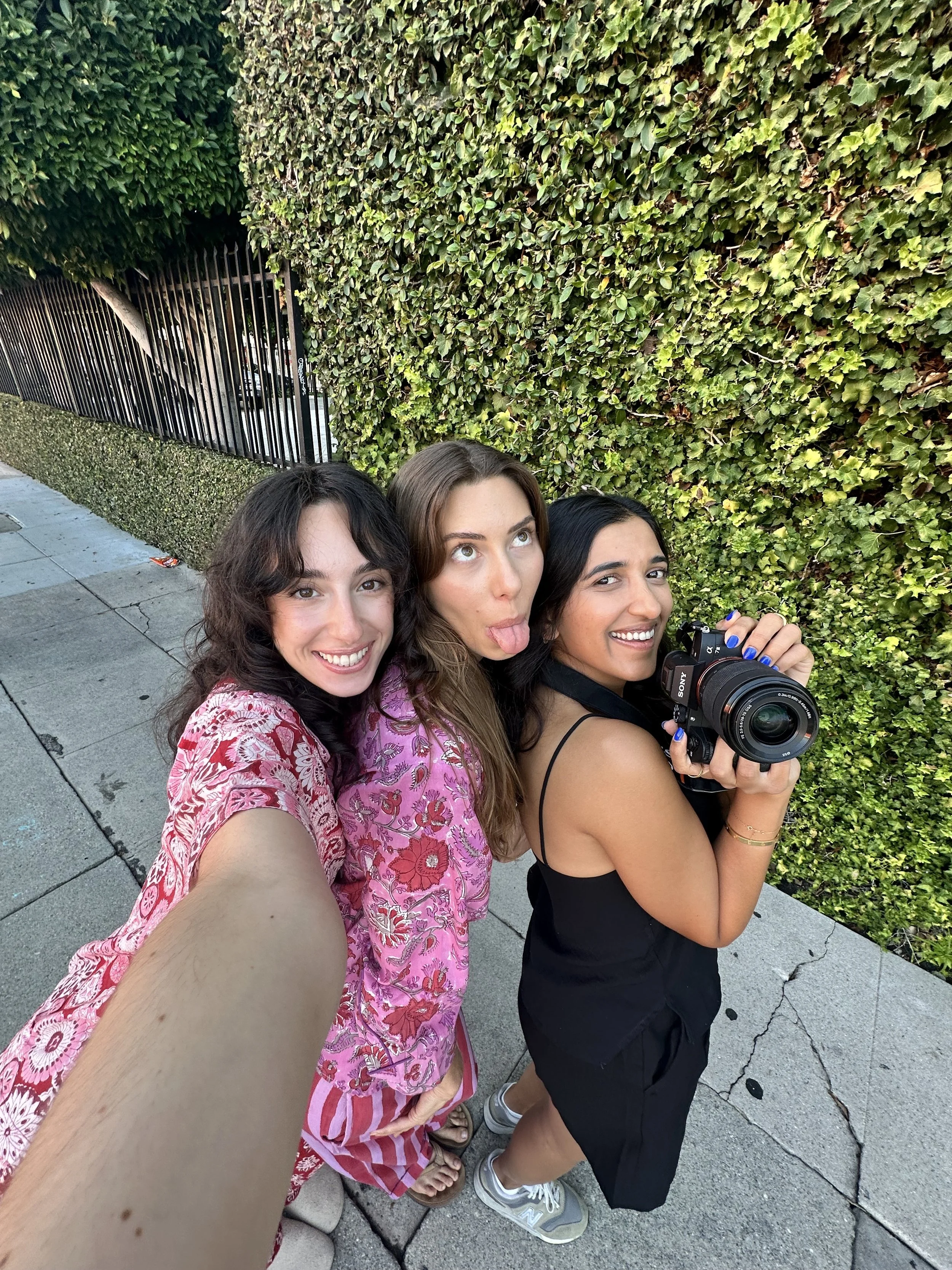 Three women taking a selfie on a sidewalk with a leafy green wall in the background.