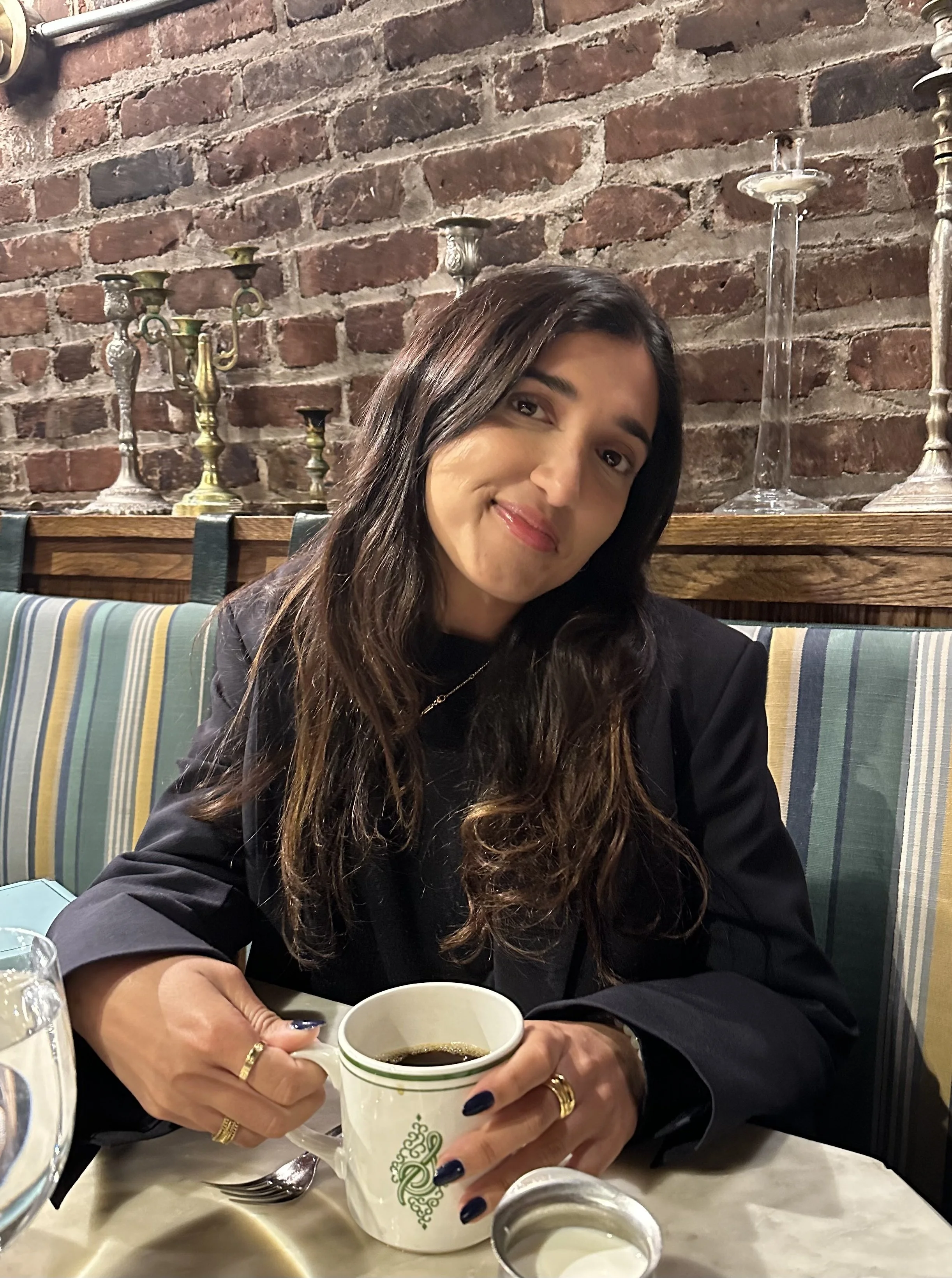 A woman with long dark hair and a black jacket sitting at a table in a restaurant, holding a cup of coffee and smiling, with a brick wall and candlestick holders in the background.