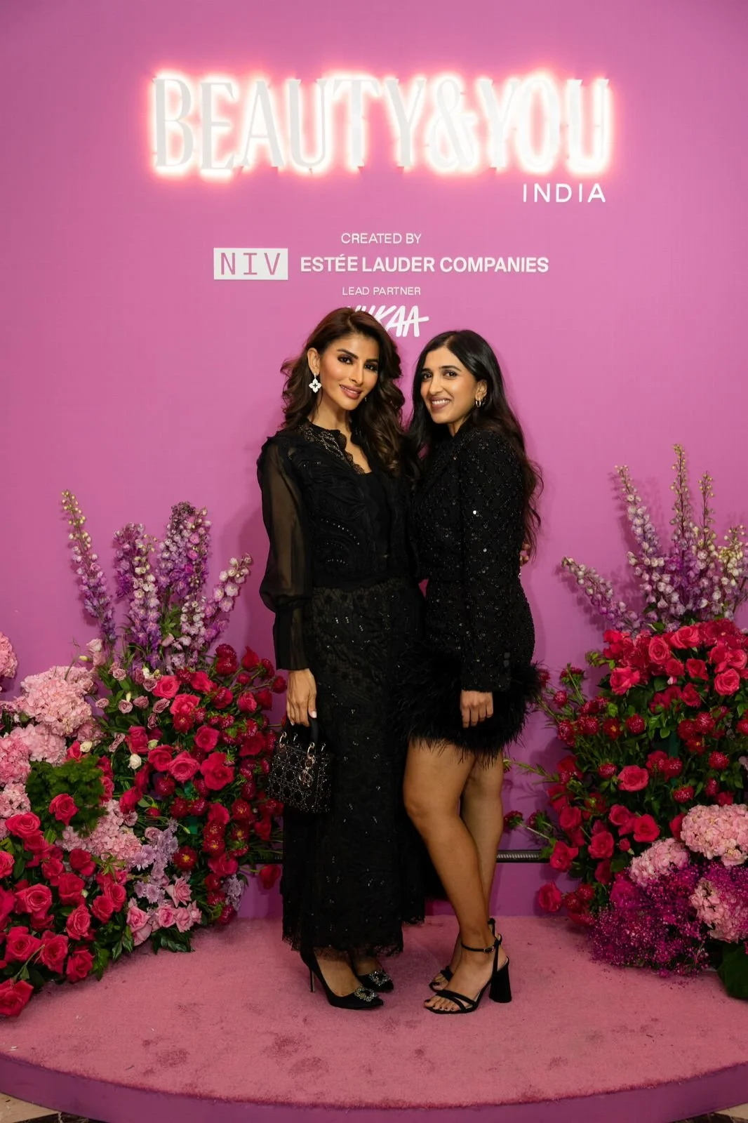 Two women dressed in black standing together on a pink carpet with a purple and pink floral background, at an event called "Beauty & You India."