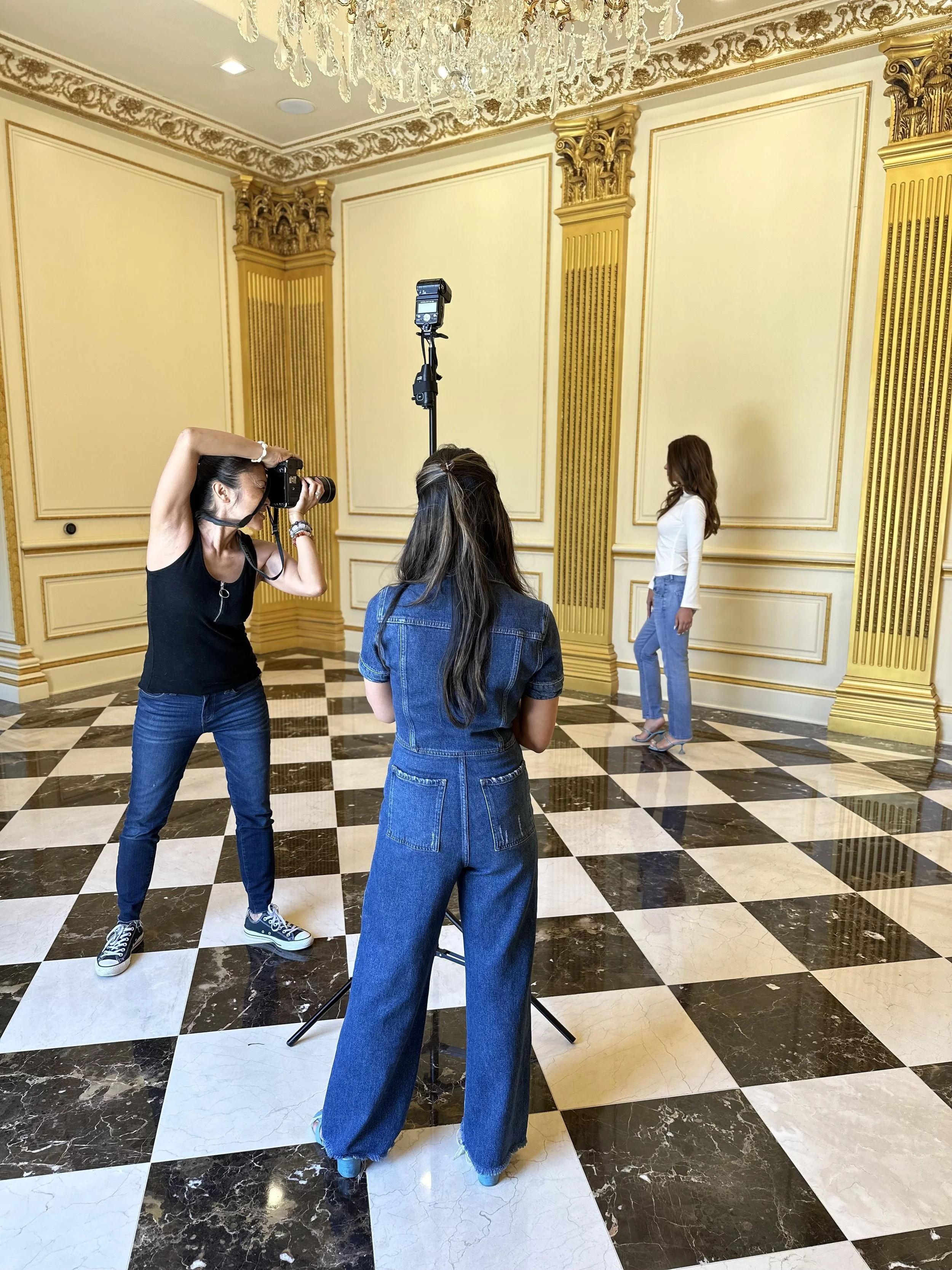 A woman is getting her portrait taken in a luxurious room with ornate gold accents on cream-colored walls and a chandelier overhead. Photographers are photographing her, one with a camera, another behind a tripod, with a checkered black and white marble floor.