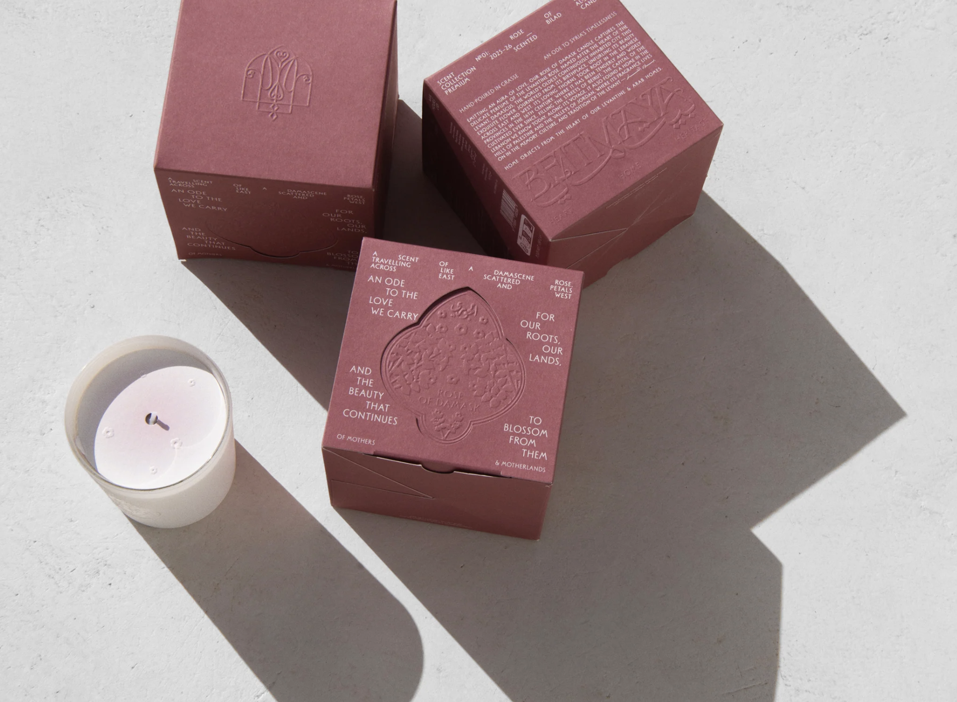 Three pink boxes labeled with floral and fragrance information, a small white candle in a glass holder, all casting long shadows on a light-colored surface.