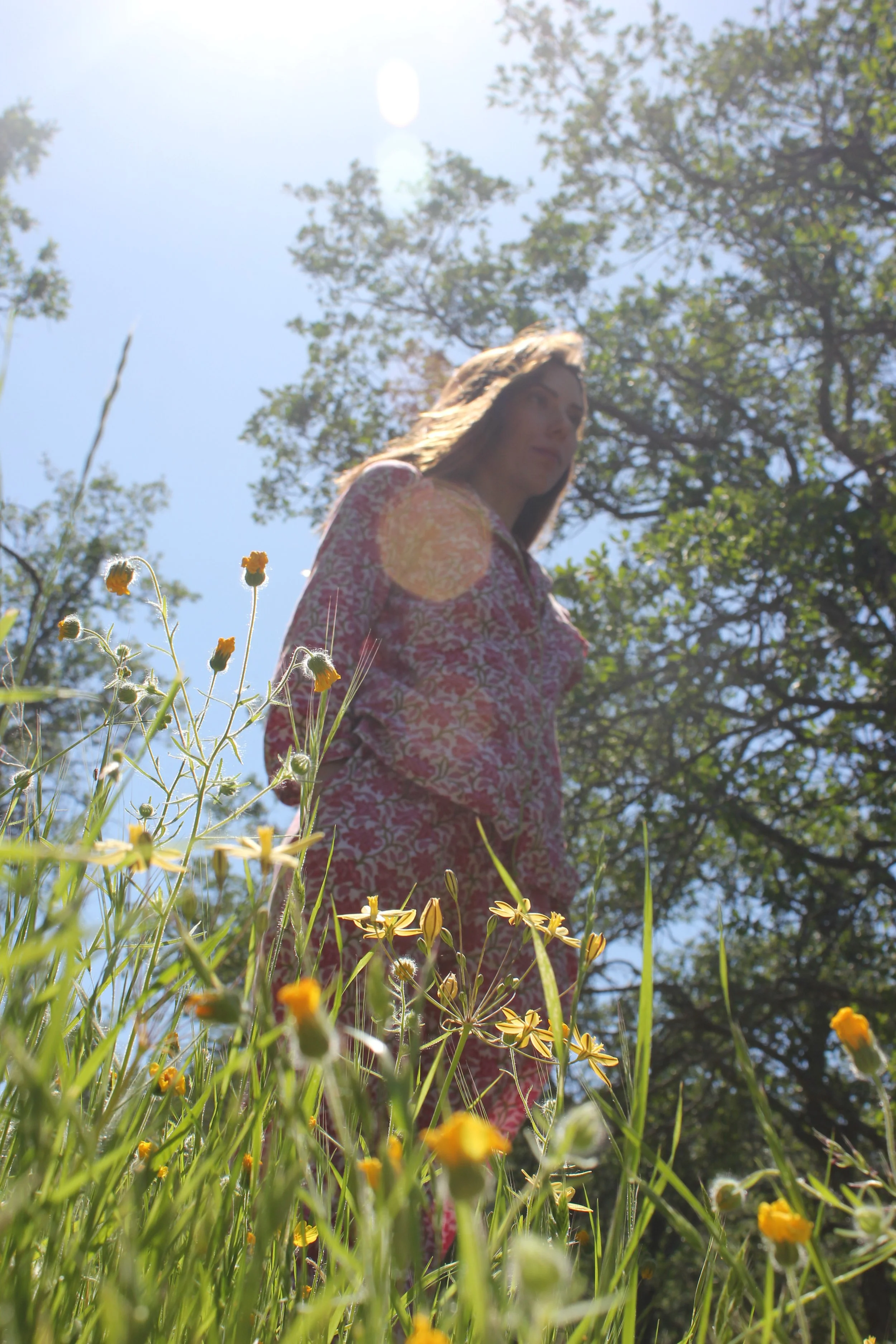 A woman standing in a field of yellow flowers with trees and blue sky in the background, sunlight creating lens flares.