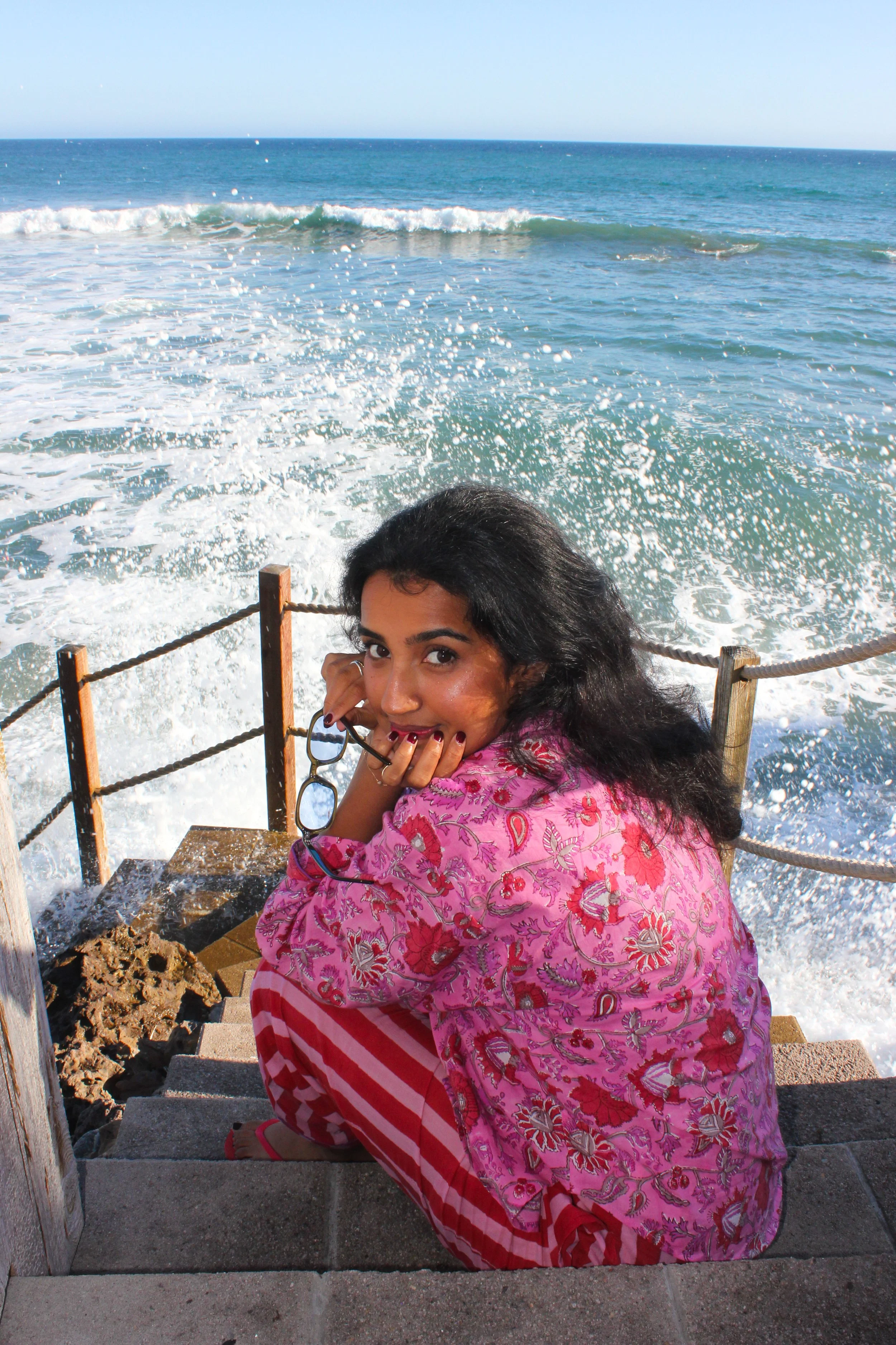 A woman in pink floral and red striped clothing is sitting on stairs near the ocean, holding glasses, with waves crashing behind her.