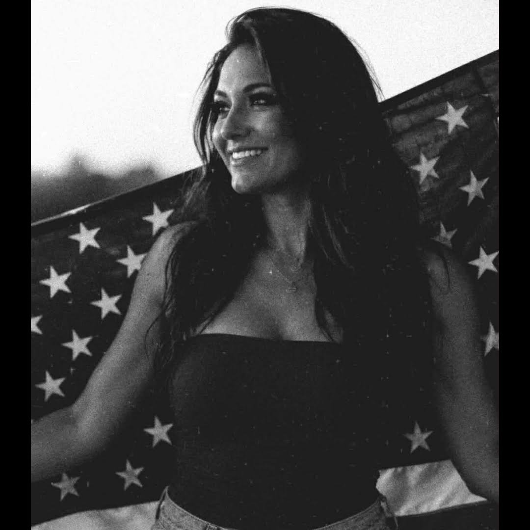 A woman with long dark hair smiling, sitting in front of an American flag background, in black and white.