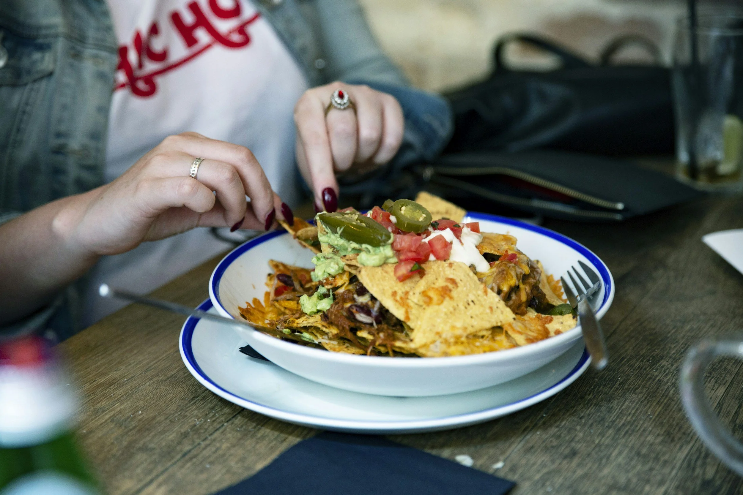 Person eating a plate of nachos topped with jalapenos, tomatoes, guacamole, sour cream, and shredded cheese at a wooden table.