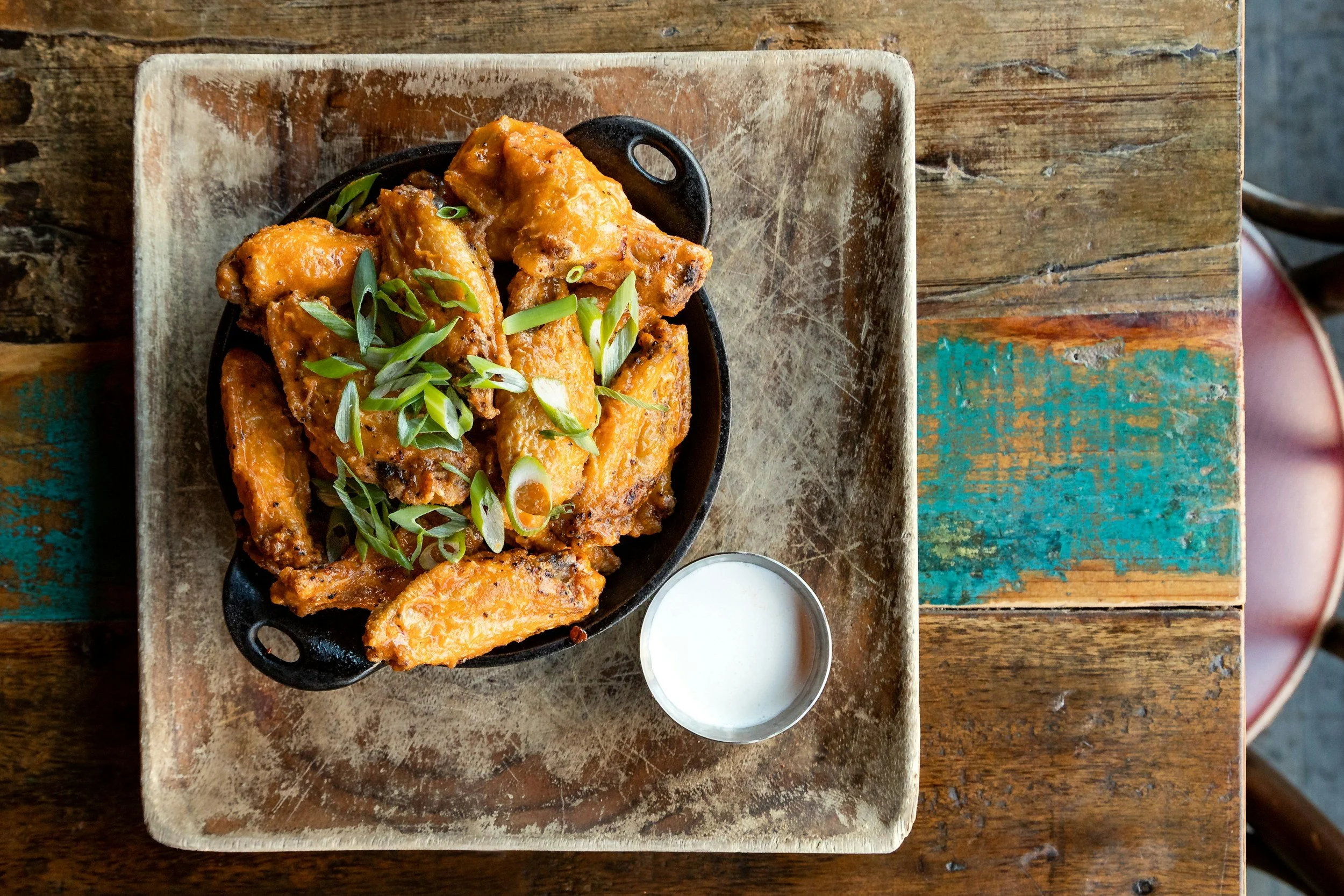 A serving of fried chicken wings garnished with sliced green onions, accompanied by a side of creamy dipping sauce on a rustic wooden table.