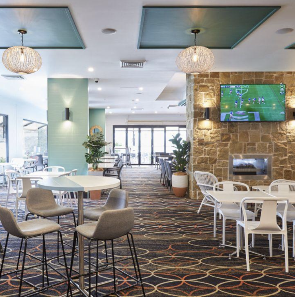 Interior of a modern lounge or restaurant with patterned carpet, white and gray chairs, tables, potted plants, a stone fireplace, and a mounted flat-screen TV showing a football game. The ceiling has decorative teal panels with hanging lights.