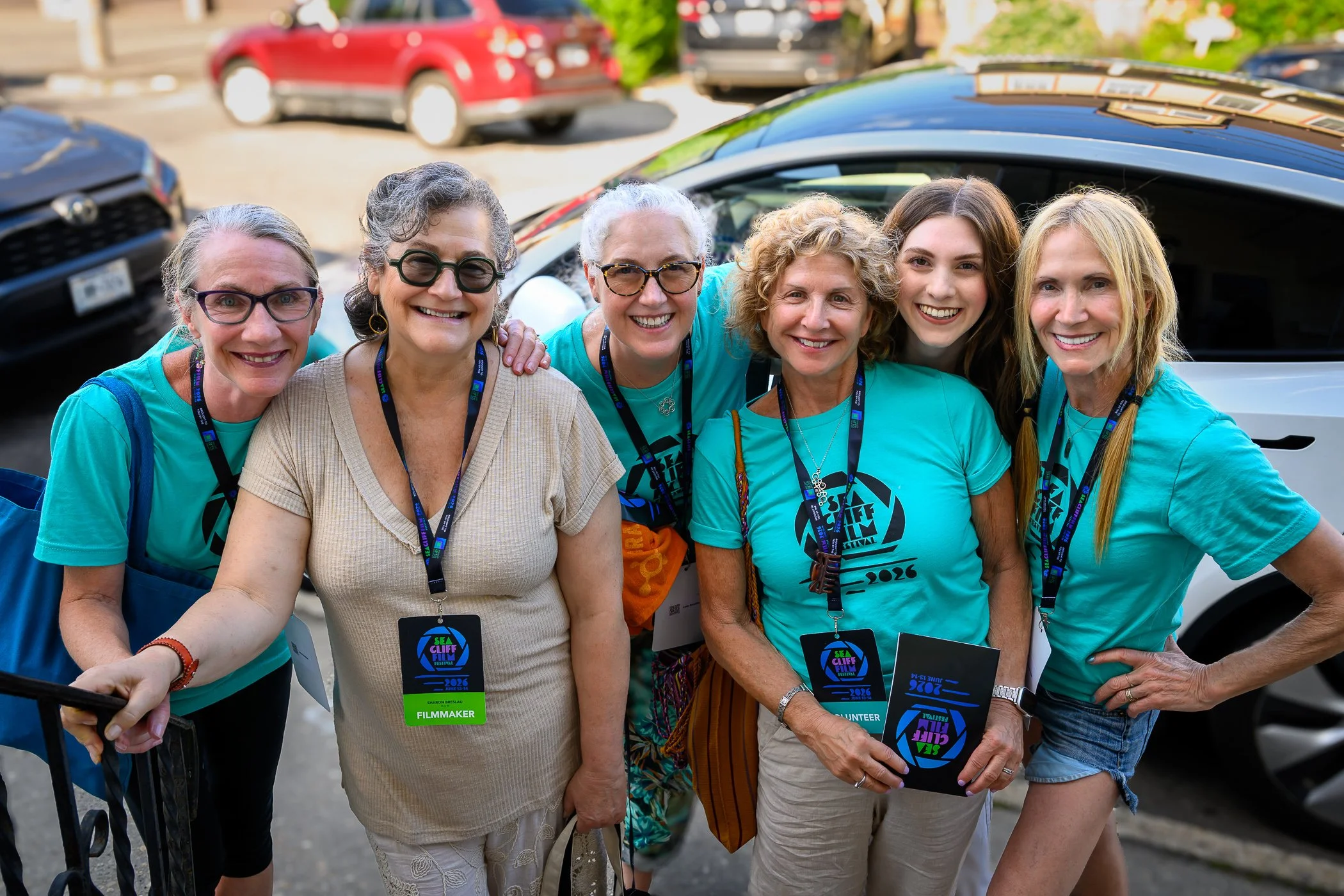 Group of seven smiling women standing close together outdoors, wearing blue t-shirts with festival logos and some with volunteer or filmmaker badges, with cars and greenery in the background.