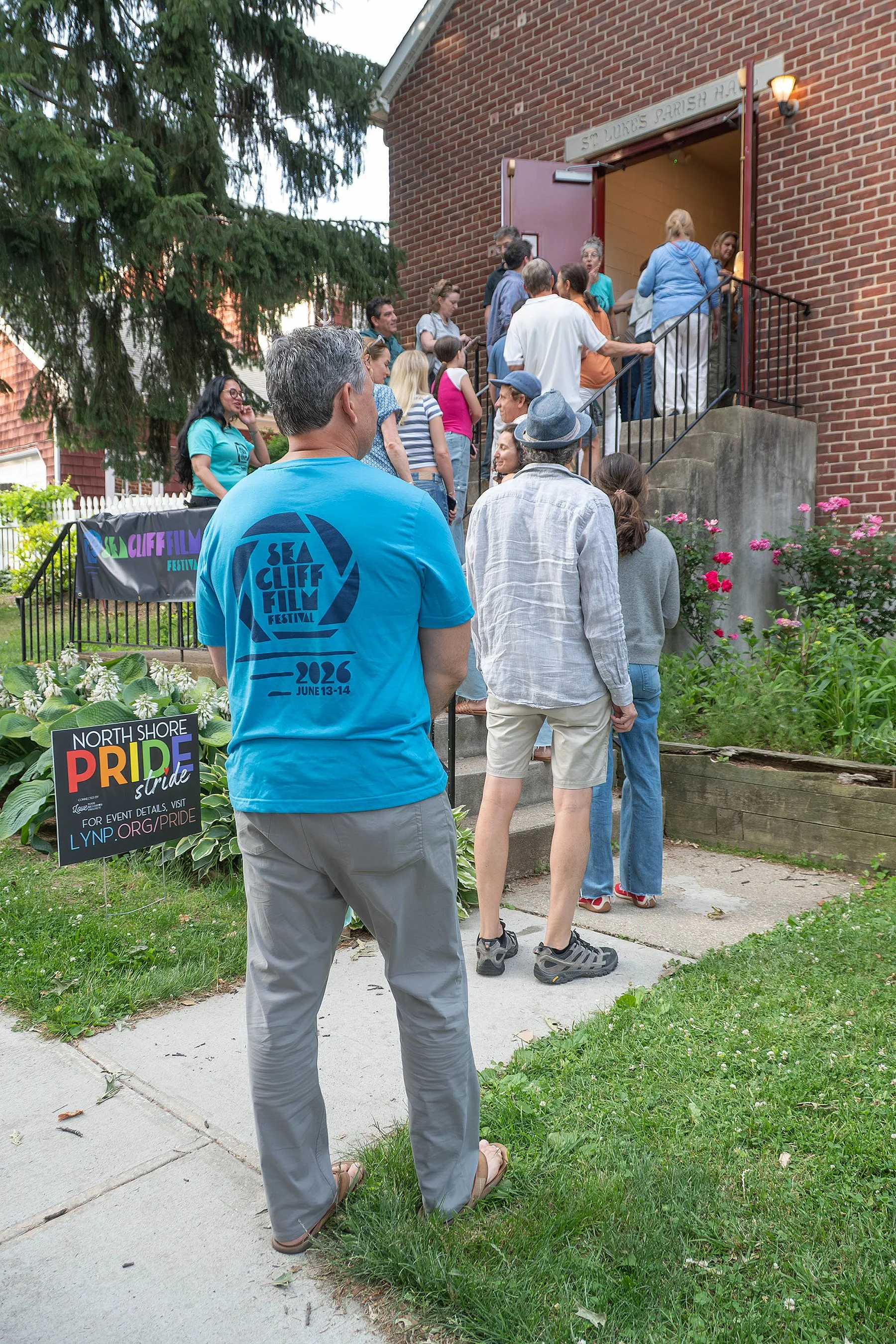 People waiting in line outside a church building with a sign for the North Shore Pride Festival, featuring a group of individuals standing and talking on the steps of the church on a residential street, with flowers and greenery in the foreground.