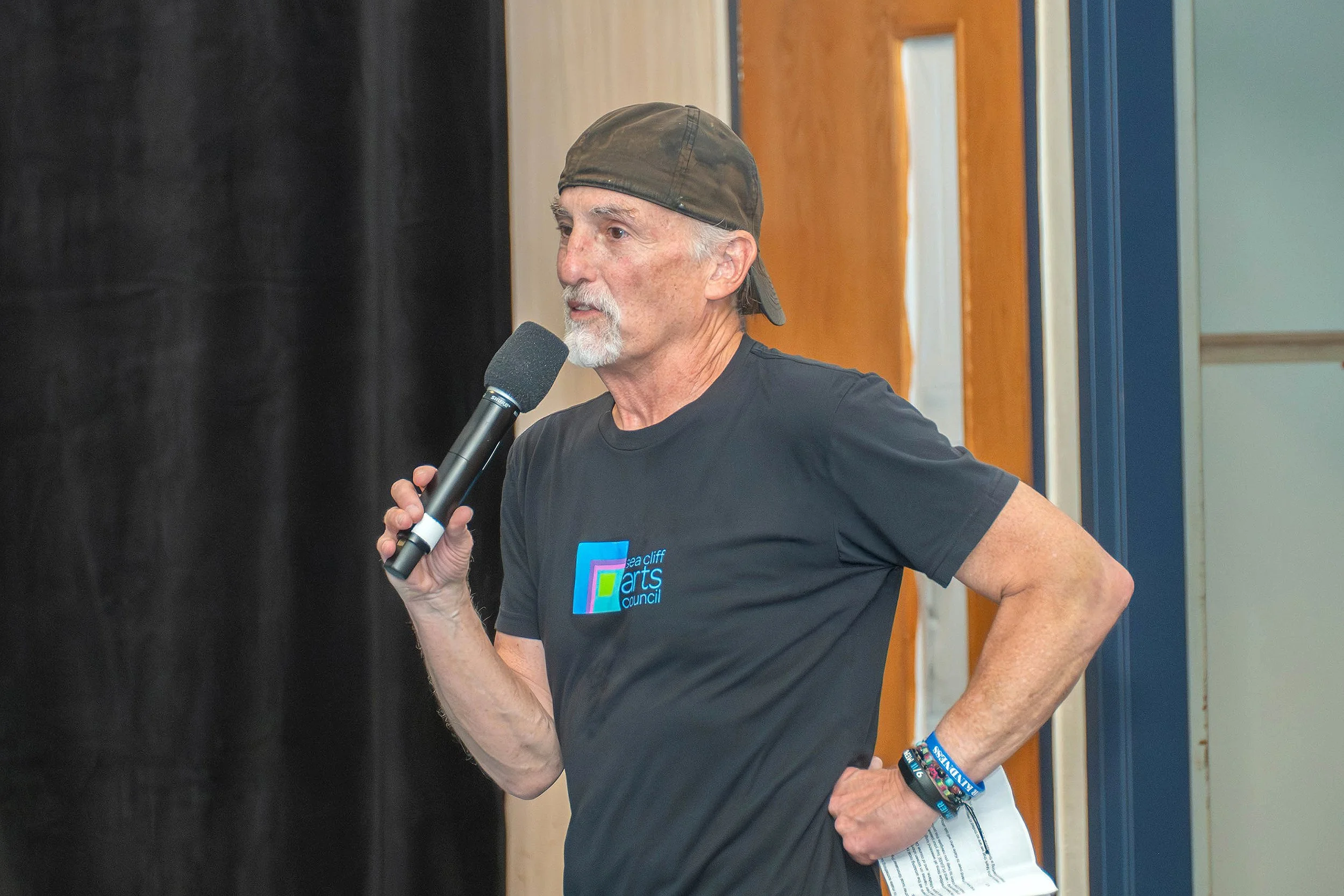 A man with gray hair and beard, wearing a brown cap backward and a black t-shirt, speaking into a microphone at an indoor event.