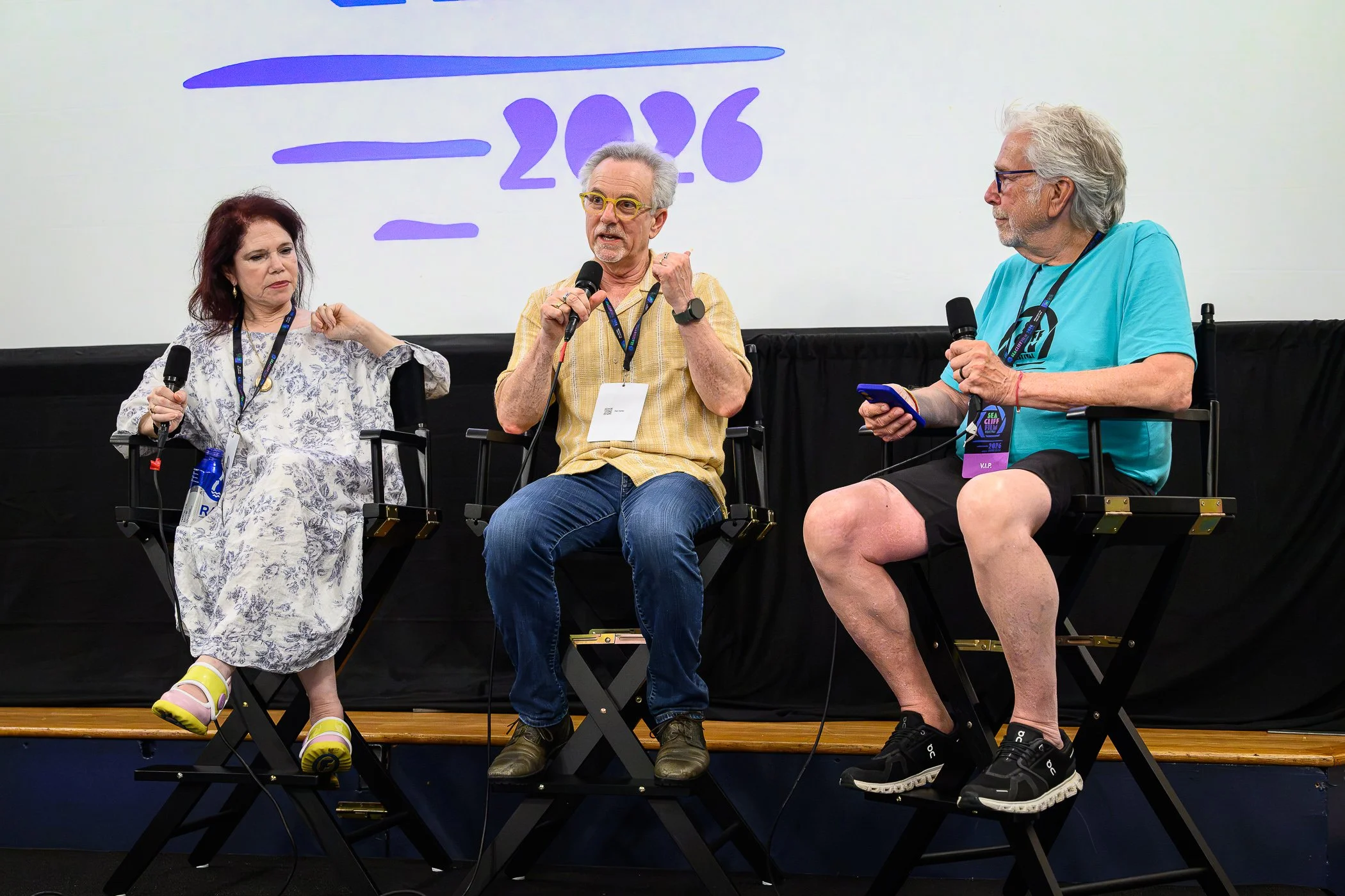 Three people seated on stage, two men and one woman, engaged in a discussion at a conference or event. The woman on the left is wearing a light-colored patterned dress and yellow sandals, the man in the middle is wearing a yellow striped shirt and glasses, and the man on the right is wearing a blue t-shirt and shorts with a badge hanging around his neck. They are holding microphones and appear to be actively participating in a panel or discussion, with a large screen behind them displaying the year '2026' and some decorative graphics.