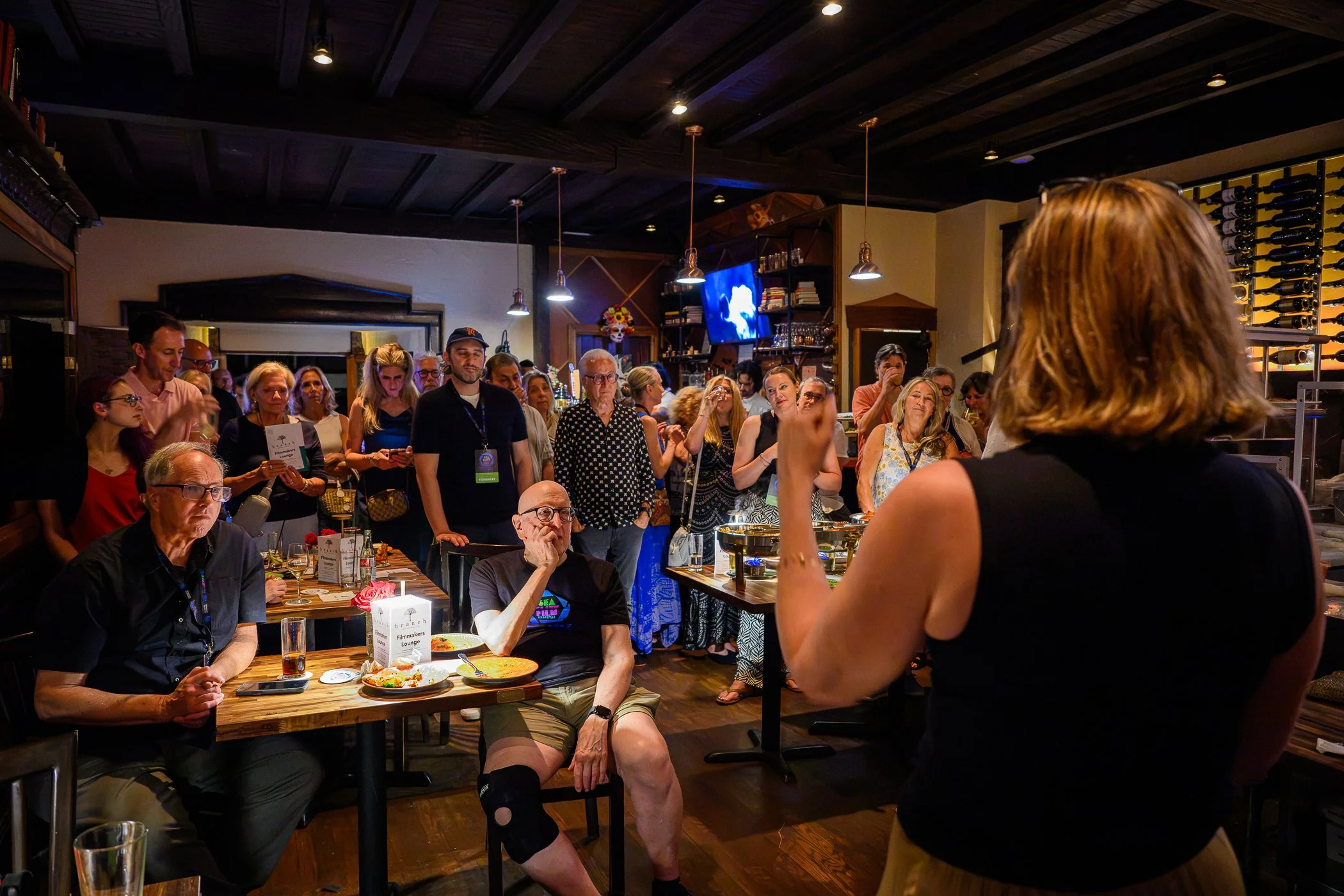 A group of people gathered in a restaurant for a presentation or talk, with a woman in the foreground speaking to the audience. The room has a cozy, rustic decor with wooden ceiling beams and shelves displaying wine bottles.
