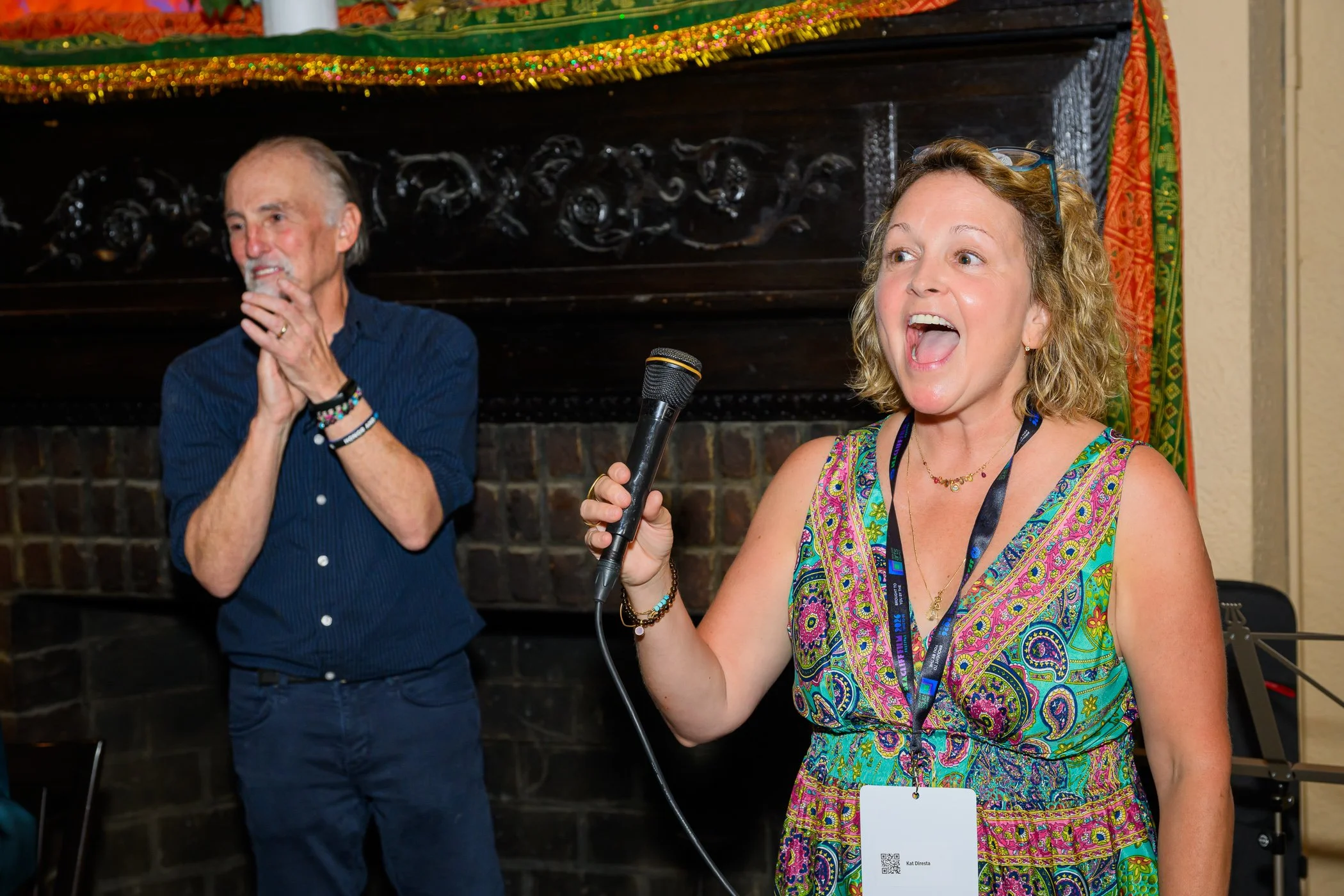 A woman with curly blonde hair, wearing a colorful paisley dress, holding a microphone, speaking or singing, with a lanyard and badge. Behind her, a man with gray hair and beard, wearing a dark blue shirt, is clapping or smiling. They are in a decorated indoor setting with a fireplace and colorful curtains.