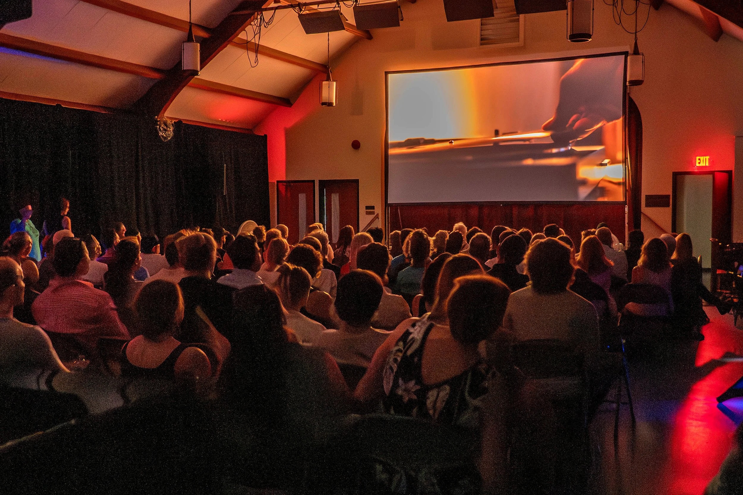 Inside a theater or auditorium with a large screen displaying a close-up of a person playing a violin. Audience members are seated and watching the screen.