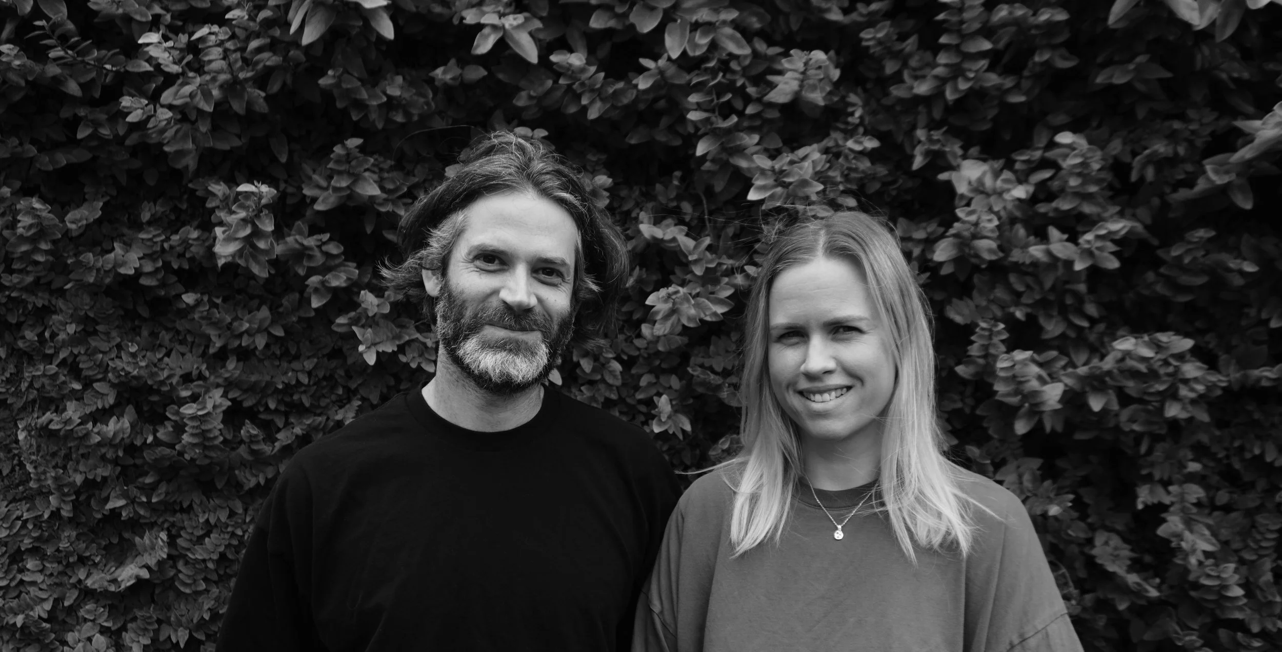 A black and white photo of a man with shoulder-length hair, beard, wearing a black shirt, and a woman with long blonde hair, wearing a casual top and a necklace with a small pendant, standing outdoors in front of a background of dense foliage, both smiling at the camera.