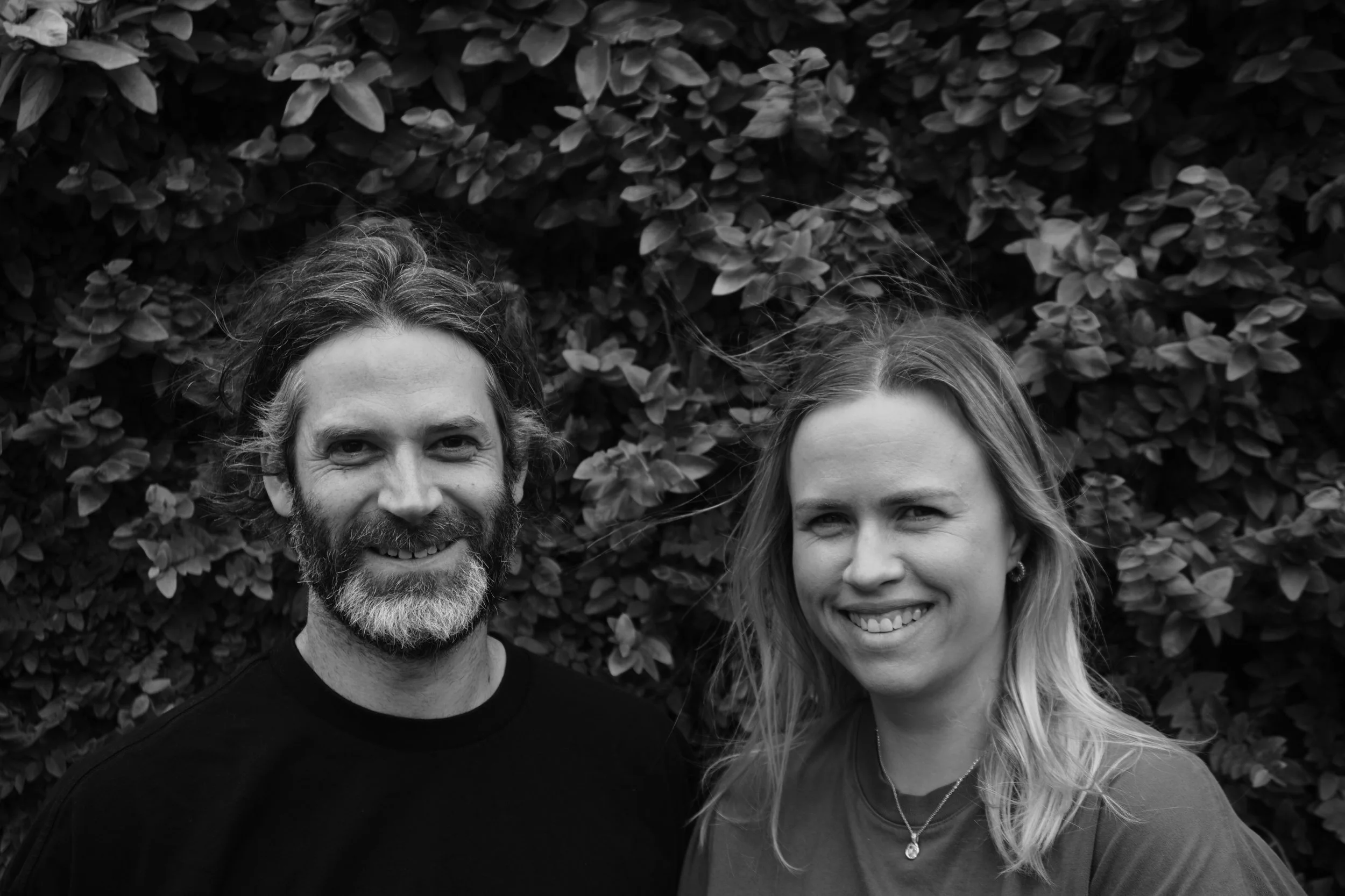 A black-and-white photo of a smiling man with a beard and a woman with wavy hair, standing in front of leafy bushes.