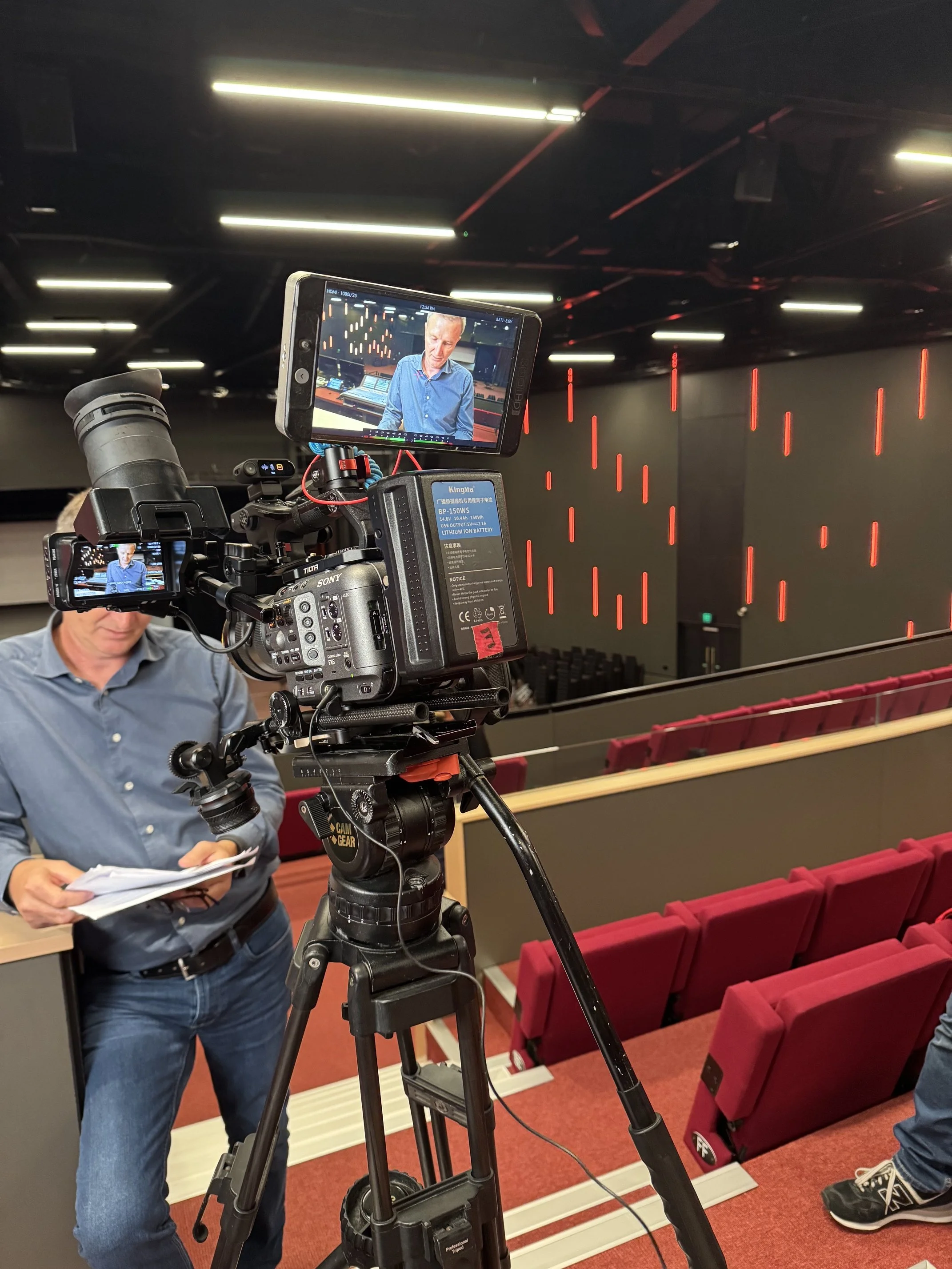 A man is being interviewed in an auditorium. A camera in the foreground shows his face