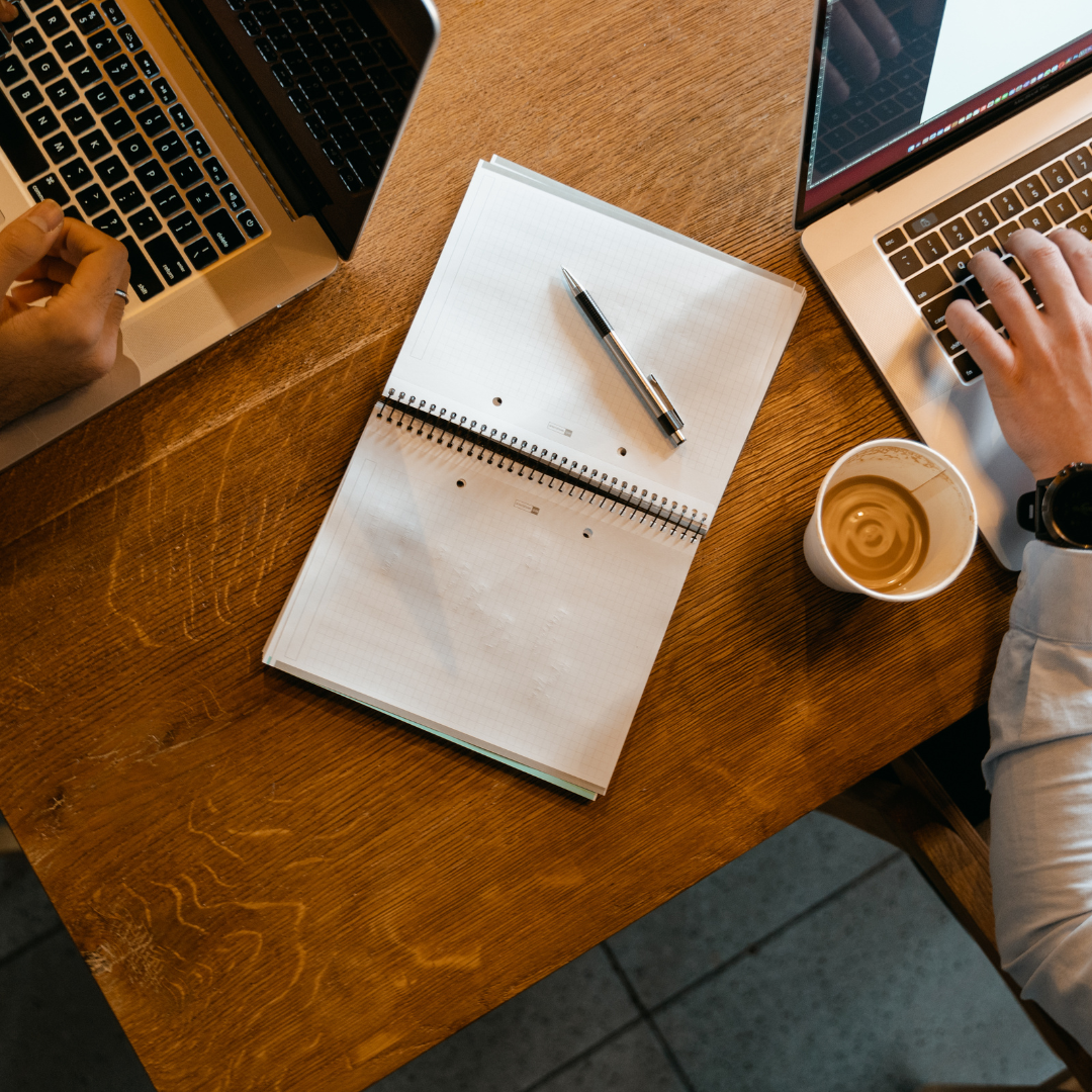 Top-down view of a wooden table with two open laptops, a spiral notebook with a pen on top, and a cup of coffee.