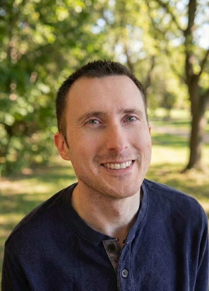 This is a portrait of a man smiling and looking at the camera. He is wearing a blue shirt with buttons and sitting in a wooded area on a sunny day.