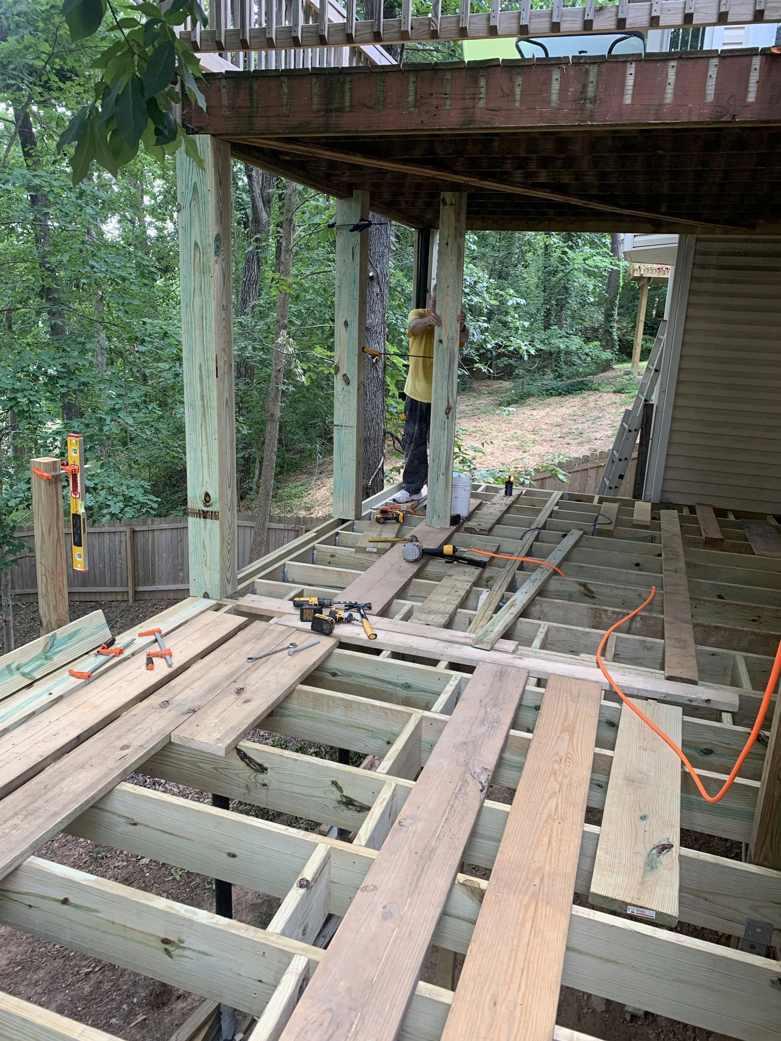 Construction workers building a wooden deck on the rear of a house in a forested area, with tools and materials scattered on the floor.