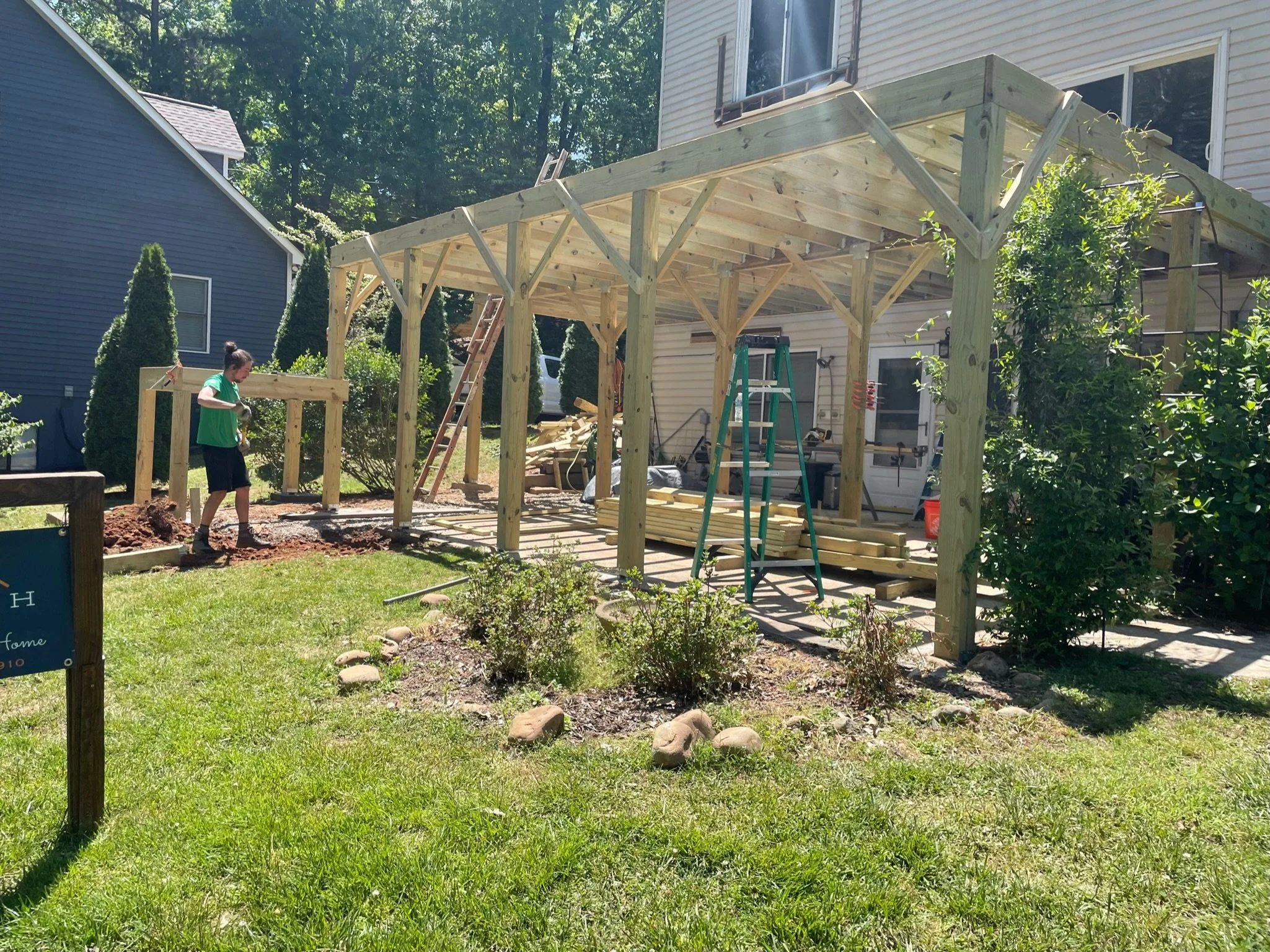 A person constructing a wooden pergola or outdoor structure in a backyard, with ladders, construction tools, and building materials present.