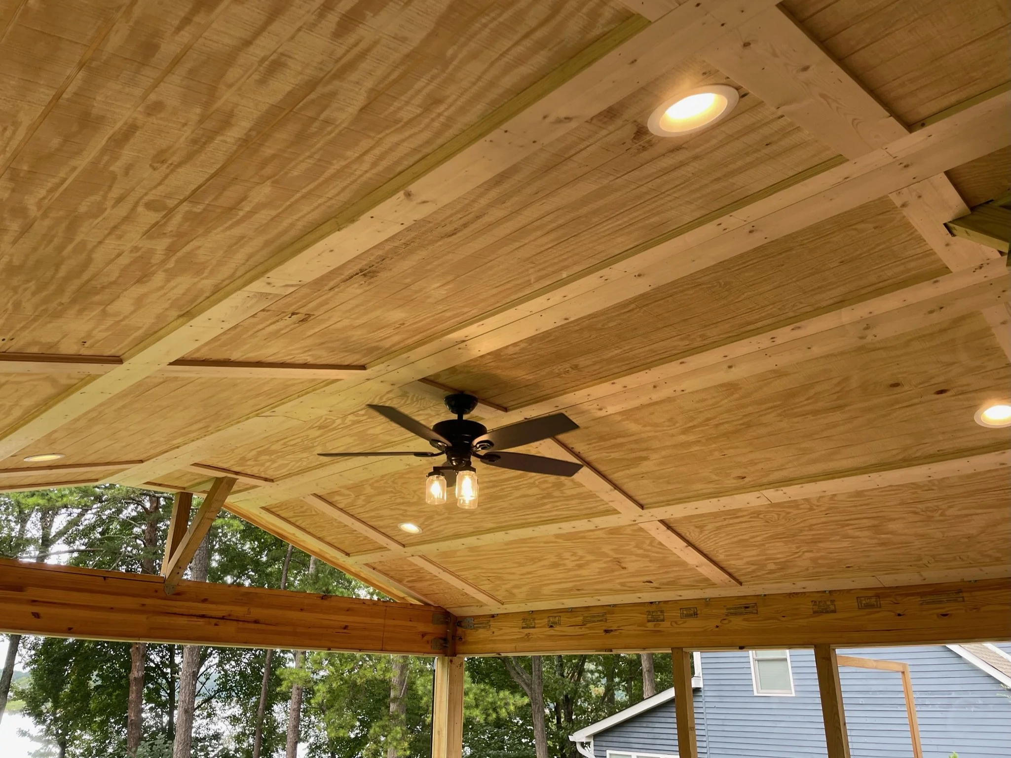 View of a partially completed wooden ceiling with recessed lighting, a ceiling fan, and outdoor trees and a neighboring house visible outside.