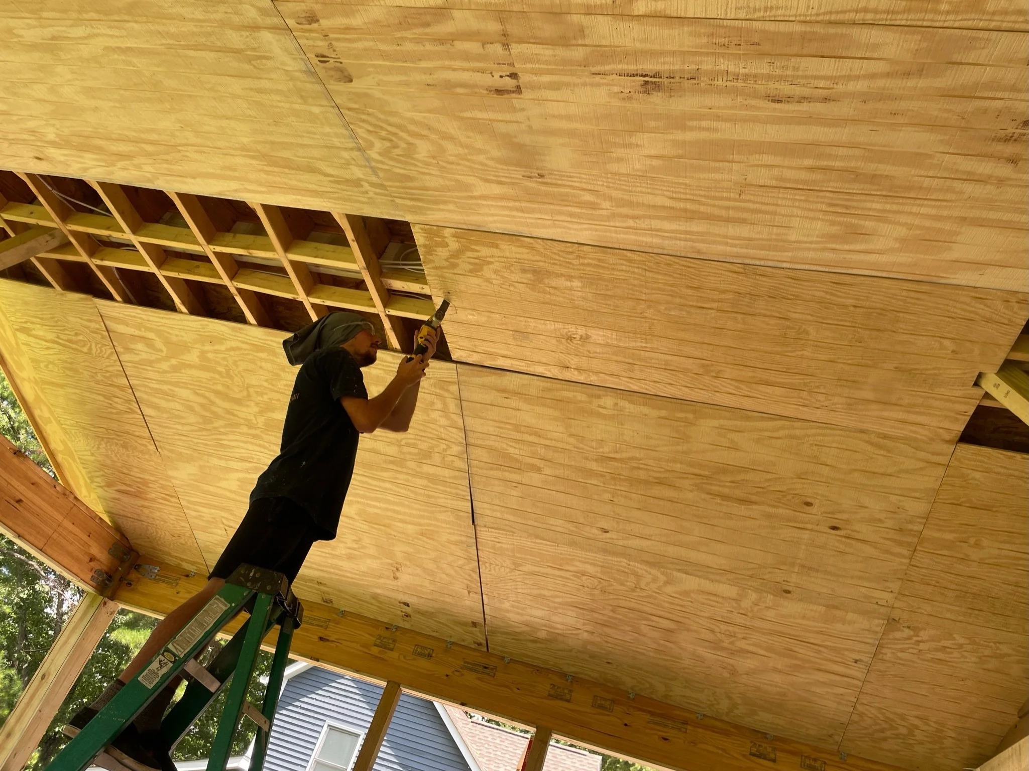 A person standing on a ladder, working on the underside of a wooden ceiling, using a power drill.