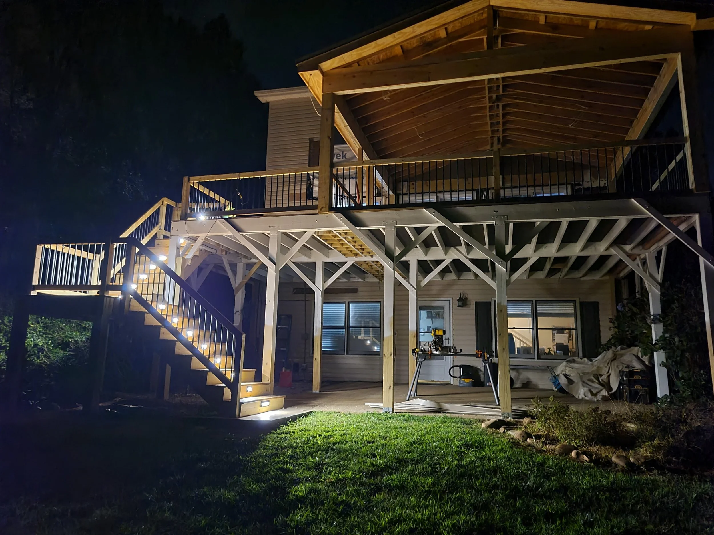 A two-story house under construction at night with illuminated stairs and interior lights visible through the windows.
