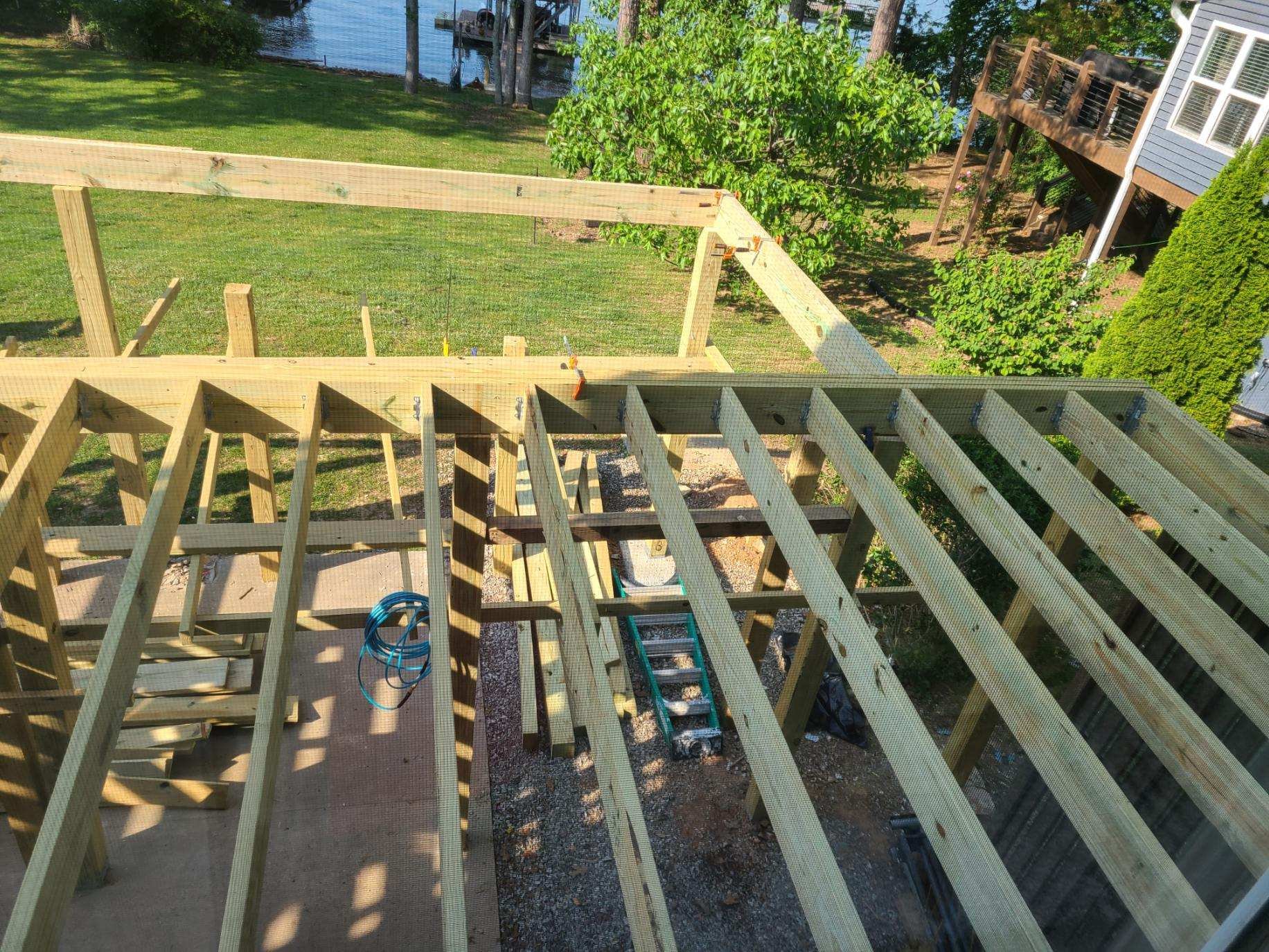 View of an unfinished wooden deck construction with framing and support beams, overlooking a green backyard and a body of water in the background.