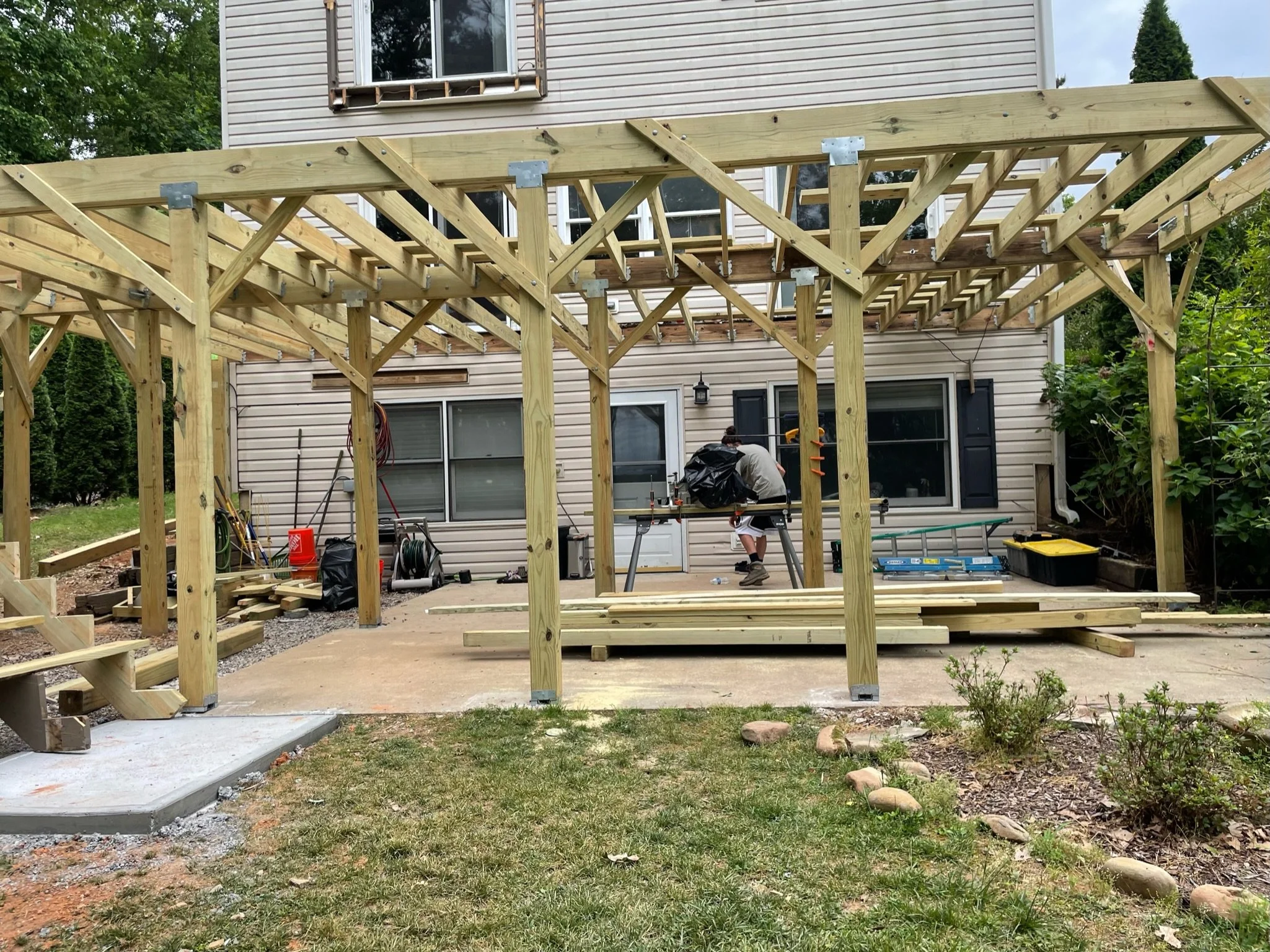 A backyard under construction with a wooden frame structure being built, a man working with power tools, and a house with beige siding and multiple windows in the background.