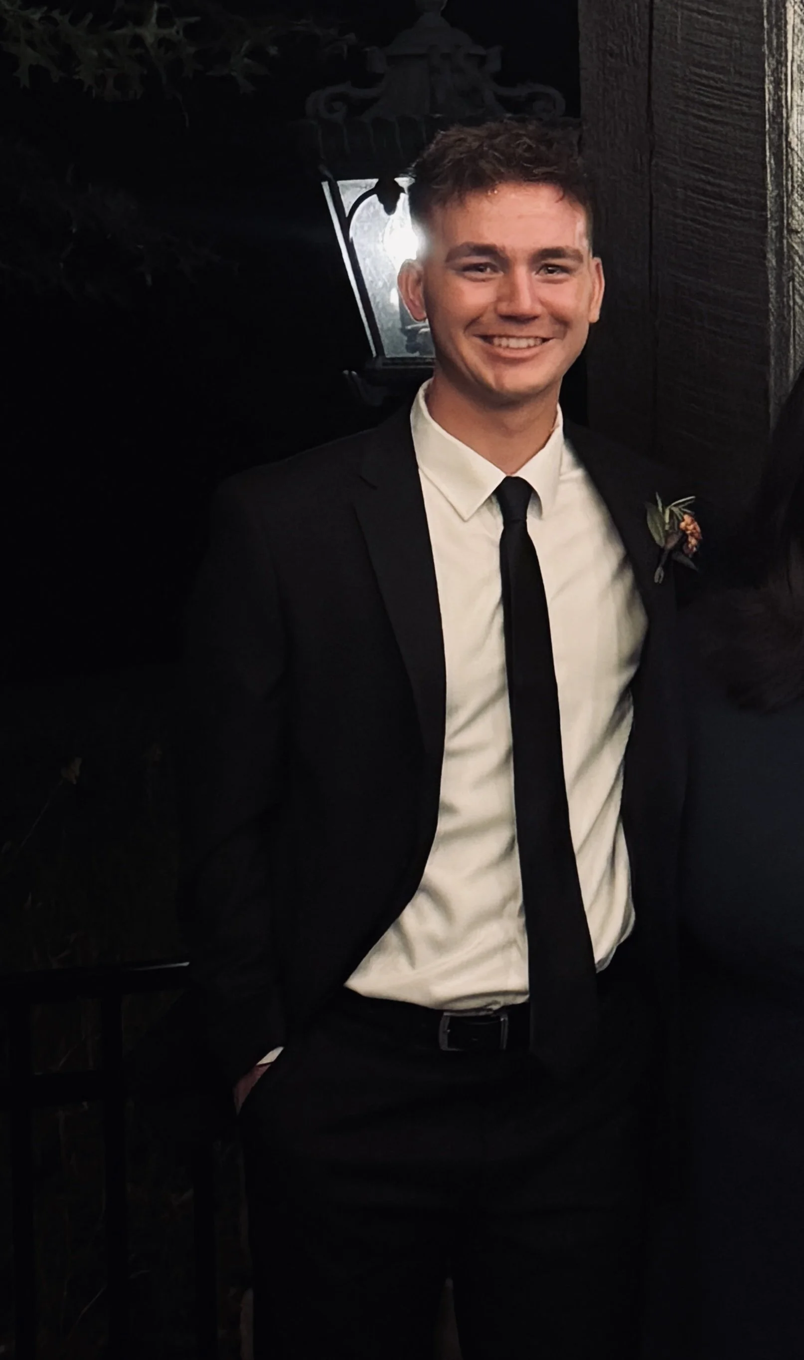 A young man in a black tuxedo with a white shirt and black tie, smiling, at a formal event at night.