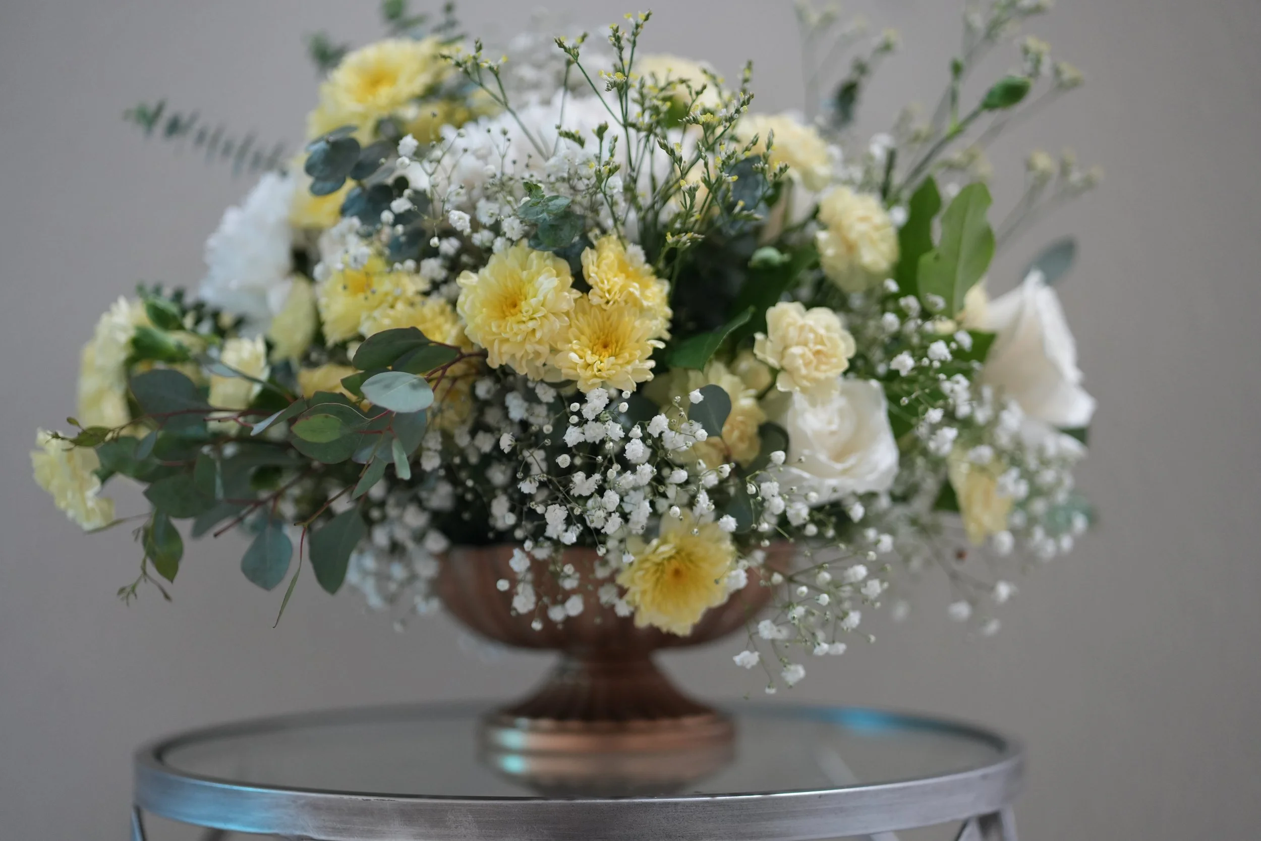 A floral arrangement with yellow carnations, white roses, baby's breath, and eucalyptus leaves in a gold ceramic vase on a silver table.