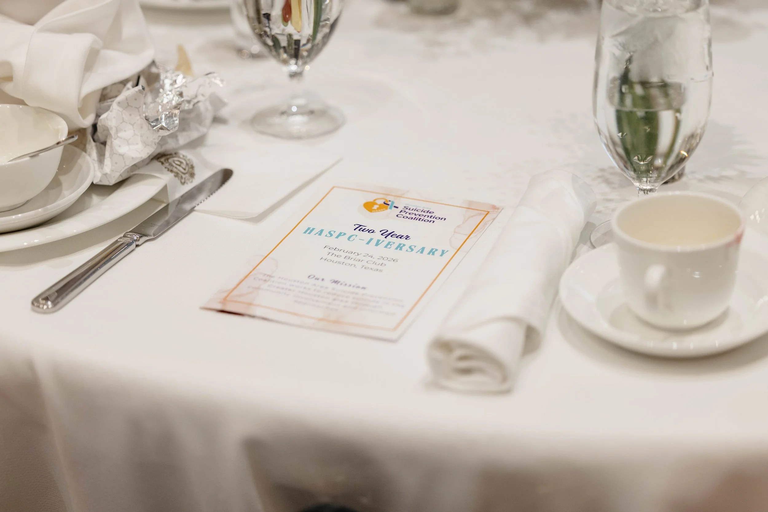 A banquet table with white tablecloth, decorated with a program that reads 'Two Year HASPC-IVERSARY February 24, 2026 The Briar Club Houston, Texas,' surrounded by glasses of water, a silver spoon and knife, white dishes, and a cluster of wrapped and