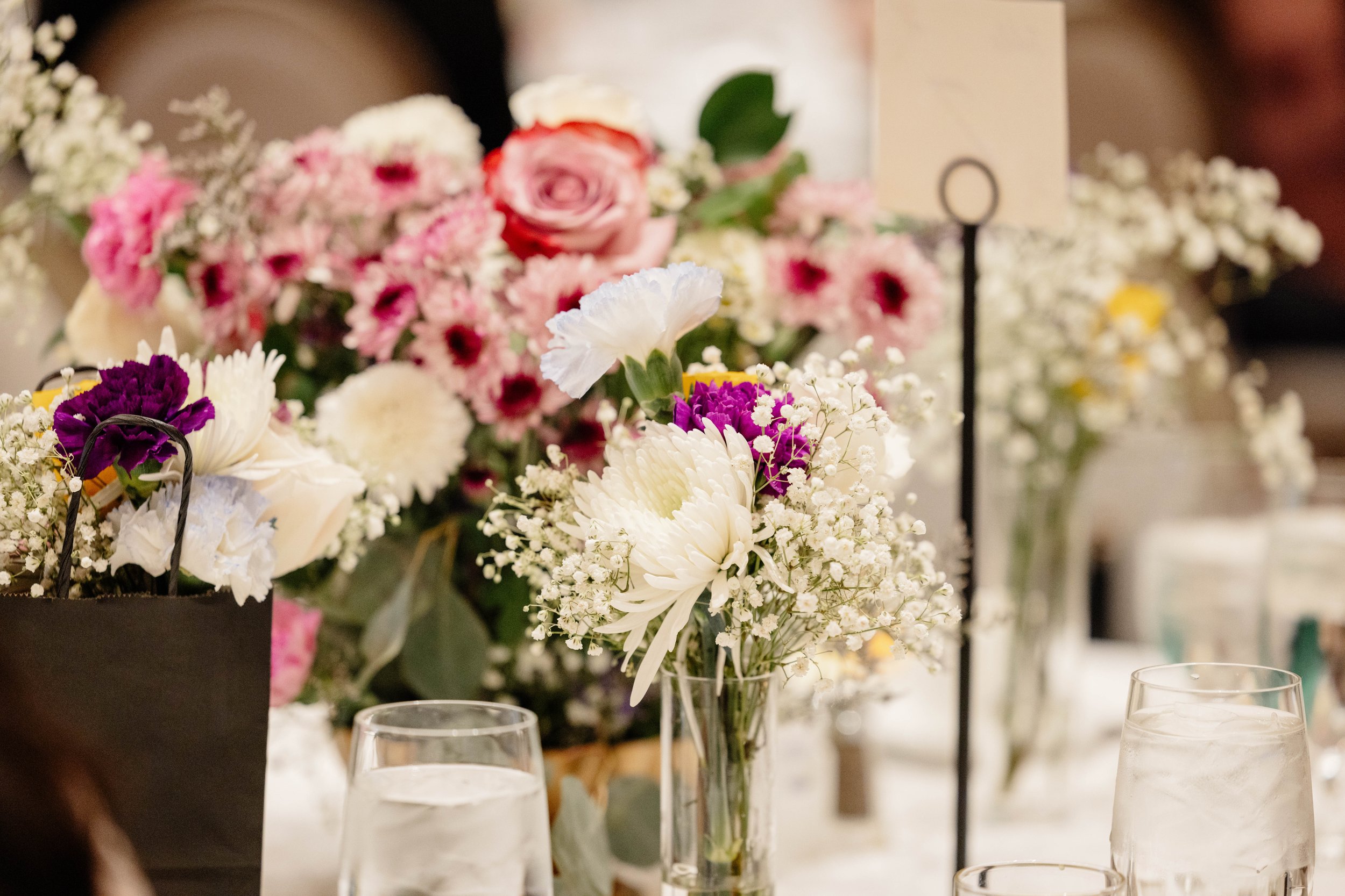 A bouquet of assorted flowers including roses, daisies, and baby's breath on a table, with glasses of water nearby, at a wedding or celebration event.