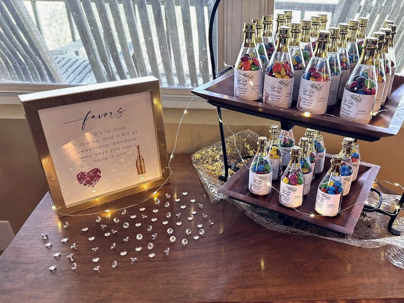 A table decorated with a lighted sign, two wooden trays with small bottles filled with colorful candies, and scattered small clear decorative stones. The sign reads: 'favors, Here's to love, bubbles, and a bit of sweetness—because who says you can't 