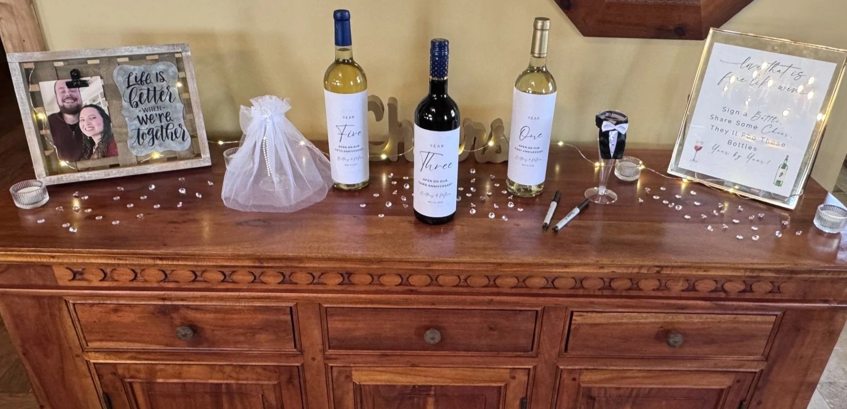 A wooden sideboard decorated for a celebration with three bottles of wine labeled for different years, a framed photo of a couple with the message 'Life is better when we're together,' a sign encouraging guests to share bottles, a black and white tux