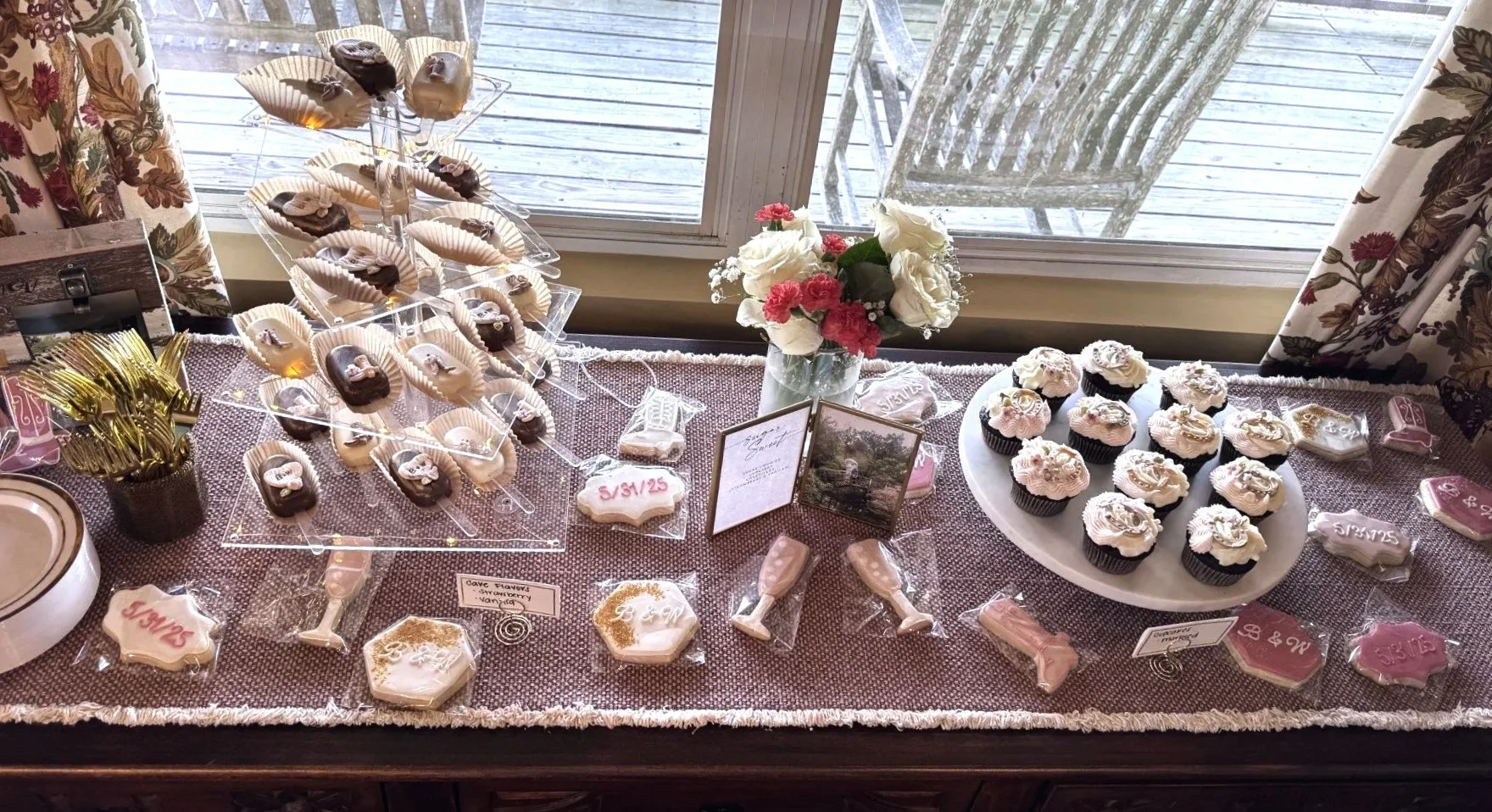 Decorative table with cupcakes, cookies, and flower arrangements for a celebration, featuring a window with outdoor view, floral curtains, and various festive treats wrapped in plastic.