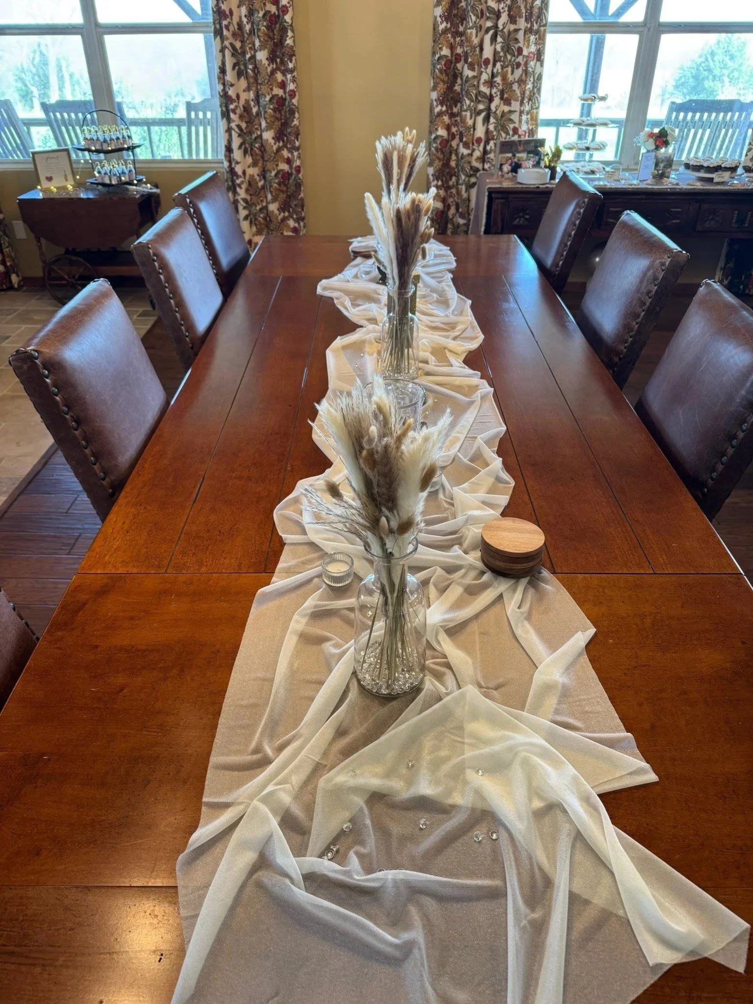 A long wooden dining table decorated with a beige fabric table runner, three glass vases filled with pampas grass, and small decorative items, set in a room with large windows and floral curtains.