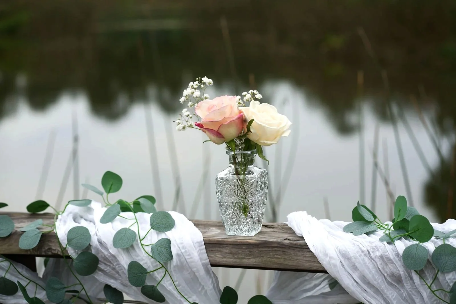 A vase with pink and white roses and baby's breath flowers on a wooden railing with greenery and white cloth, overlooking a pond.