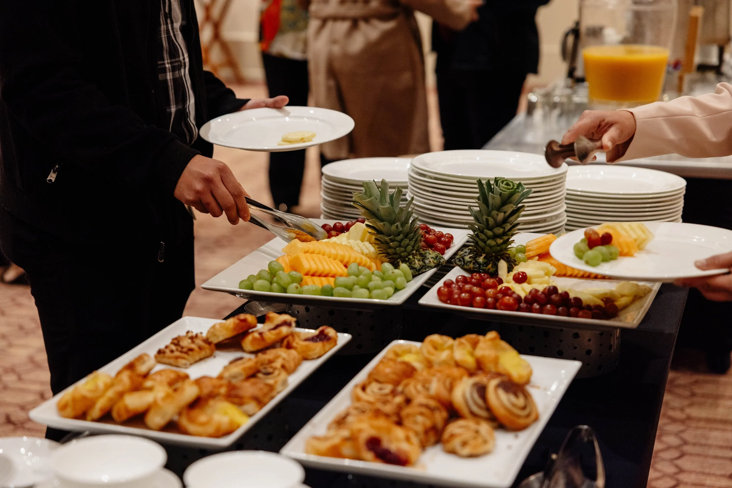 People serving themselves food at a buffet table with assorted fruit, pastries, and desserts.