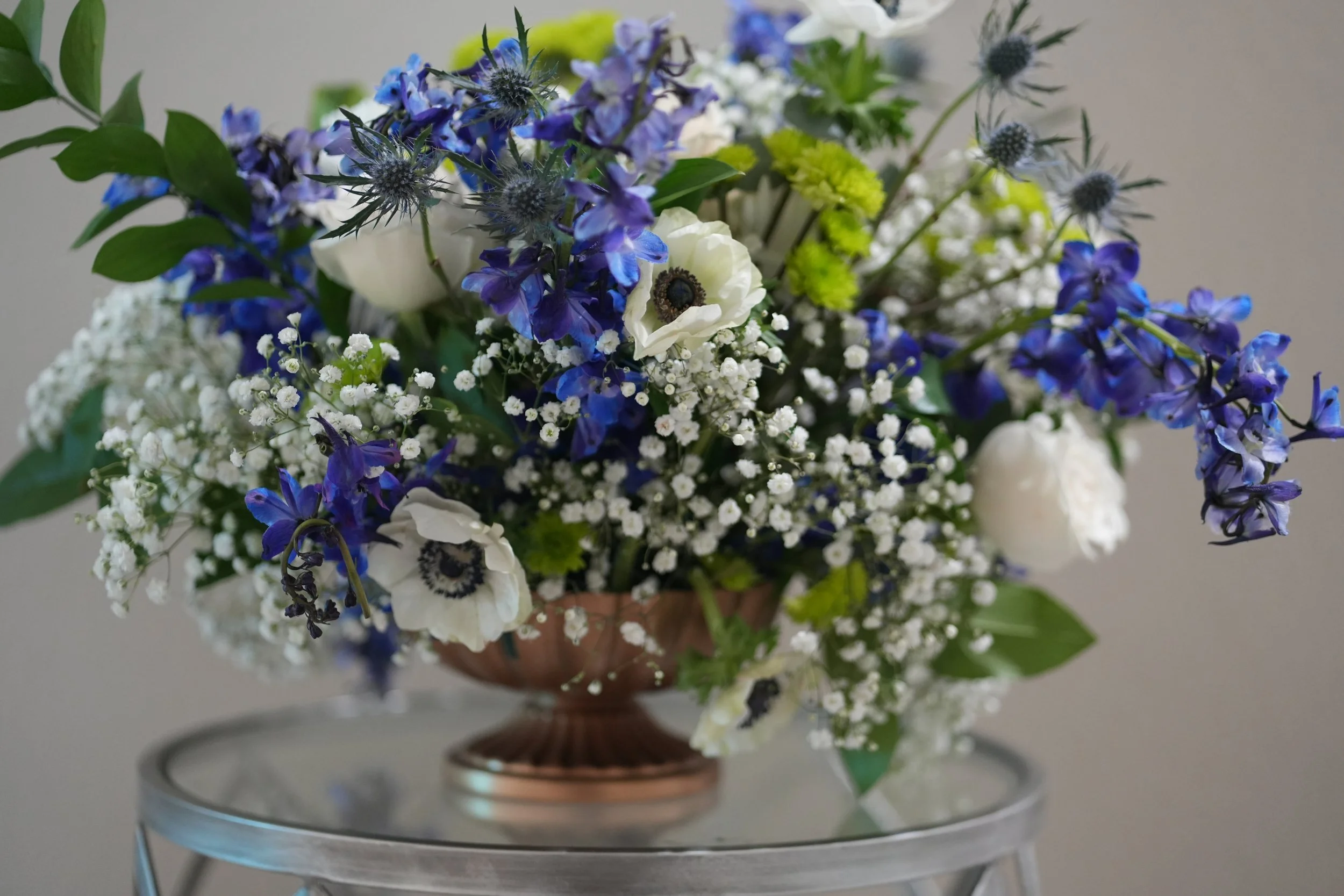 A floral arrangement with white, purple, and blue flowers, including anemones, delphiniums, baby's breath, and greenery in a gold bowl on a glass table.