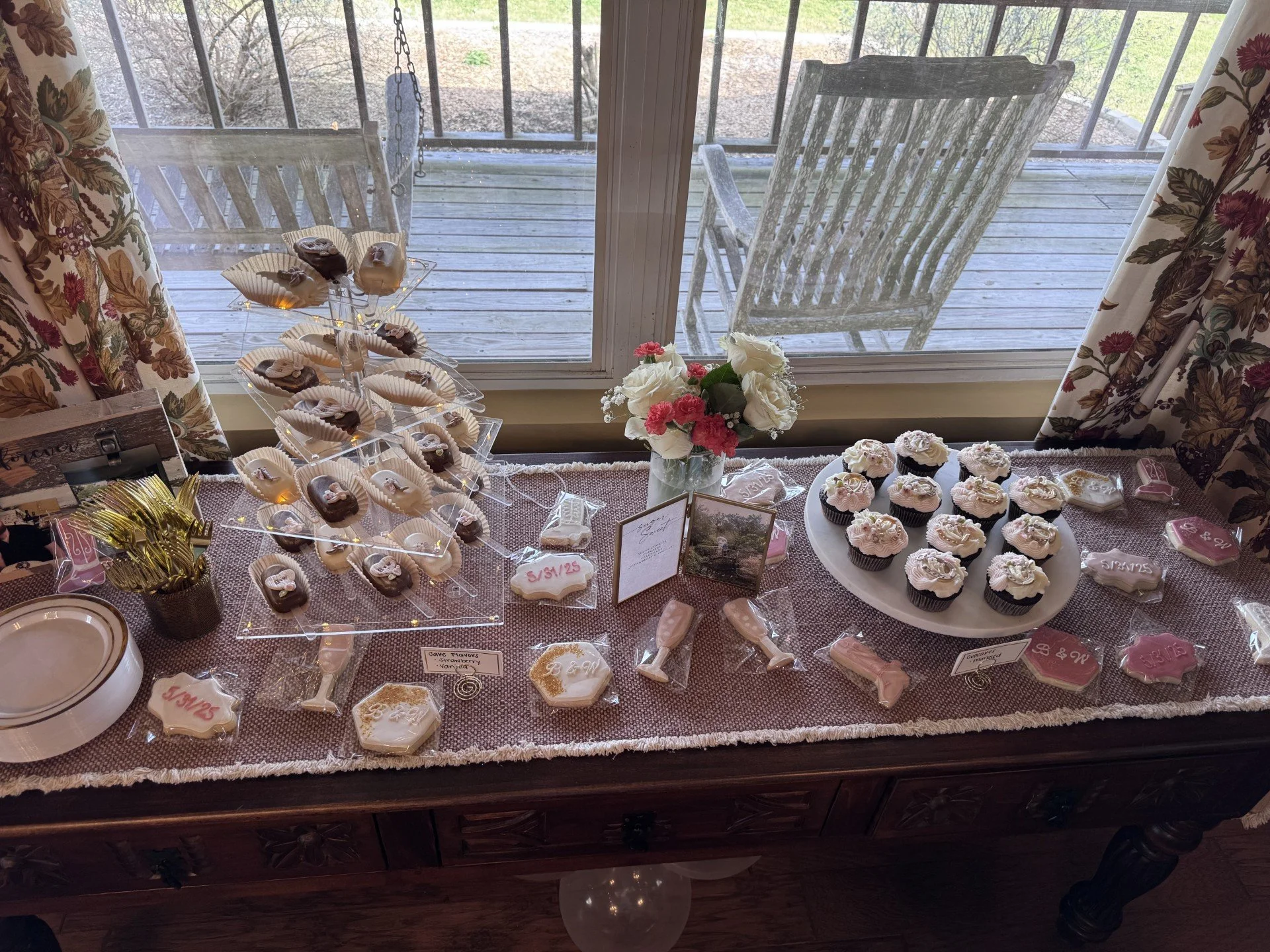 Cookies and cupcakes on a lace-covered table next to a window. Outdoor wooden deck and chairs are visible through the window. The table features decorated cookies, cupcakes with white icing, a flower arrangement, and small signs.