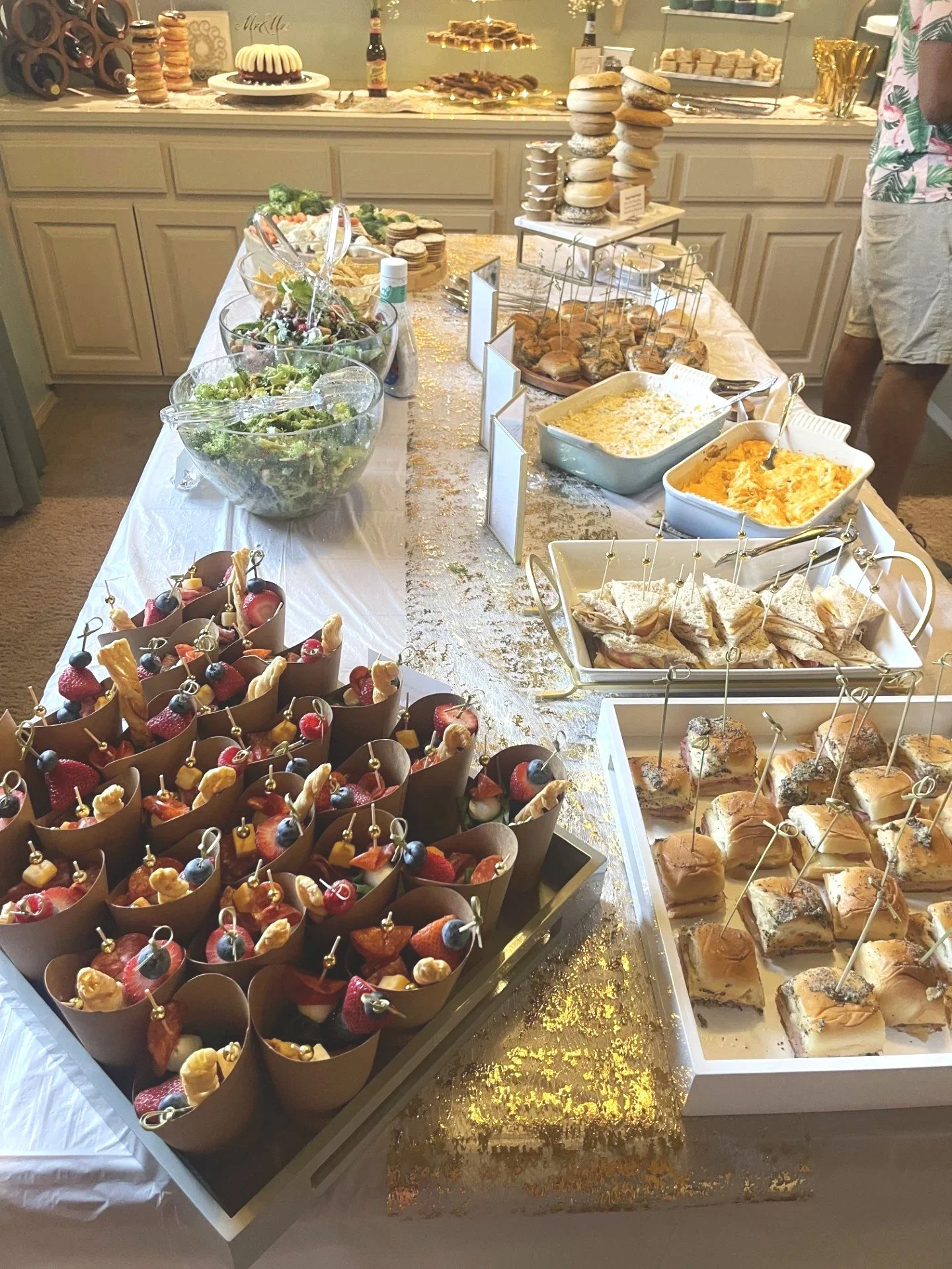 A dessert table with a variety of food and drinks, including fresh salads in large glass bowls, a bowl of cookies, a layered cake, and stacks of macarons and pastries, decorated with gold accents and set in a well-lit room.
