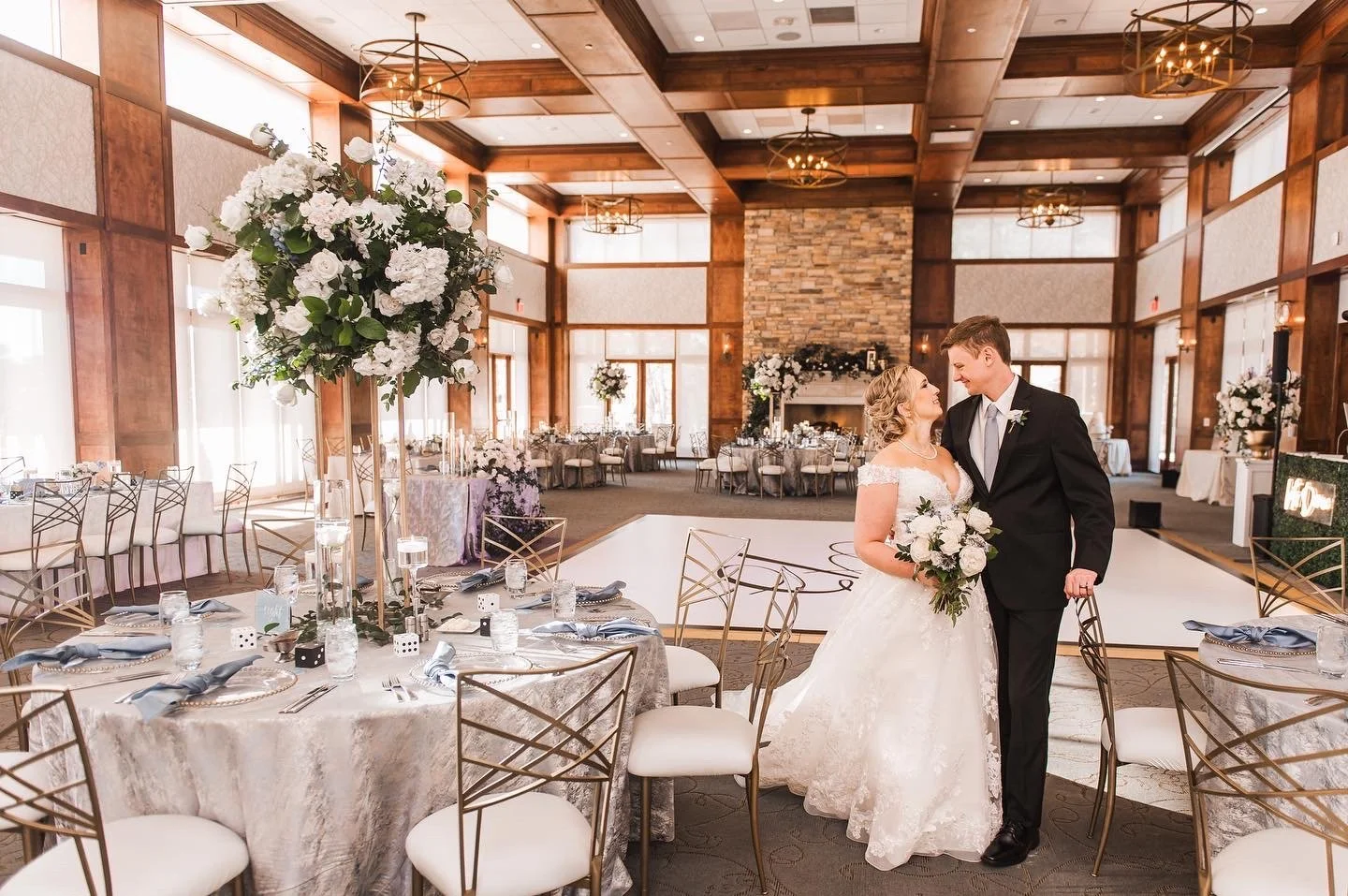 A bride and groom standing close together in a decorated wedding reception hall, smiling and looking into each other's eyes, with the bride holding a bouquet of white flowers.