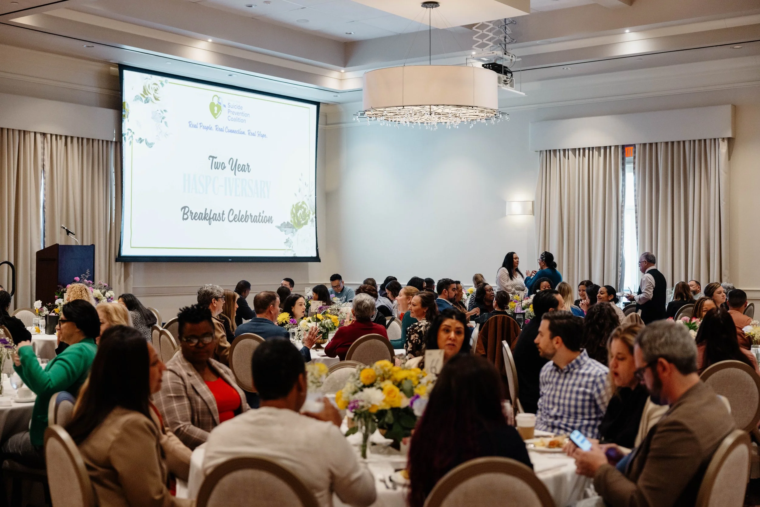A large banquet hall filled with round tables and many guests attending a two-year anniversary breakfast celebration for the Suicide Prevention Coalition. The room has a large projection screen displaying event details, floral centerpieces on each ta